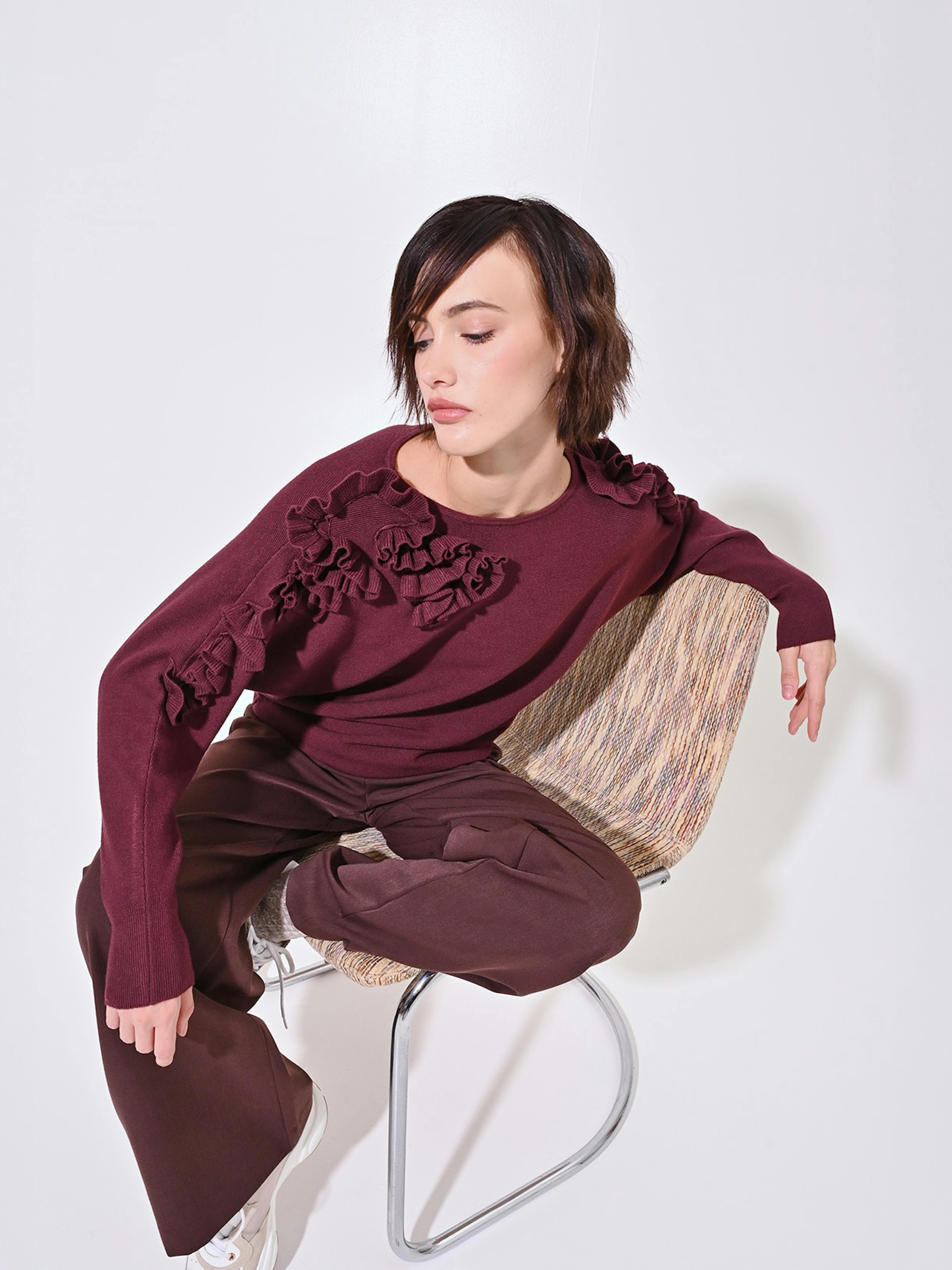 A person in a burgundy sweater with floral patterns sits on a modern chair against a white background, conveying a calm and contemplative mood.