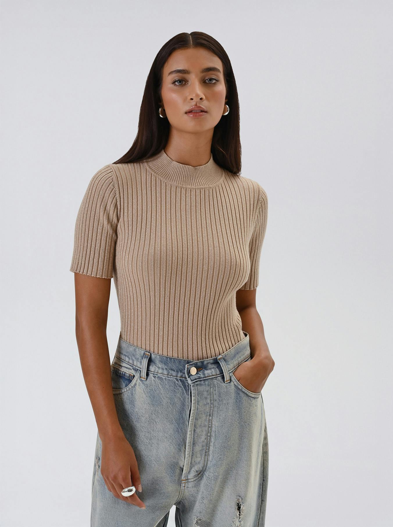 Woman in a beige ribbed sweater and light blue jeans with one hand in pocket. She's wearing silver hoop earrings, exuding a calm and confident vibe.