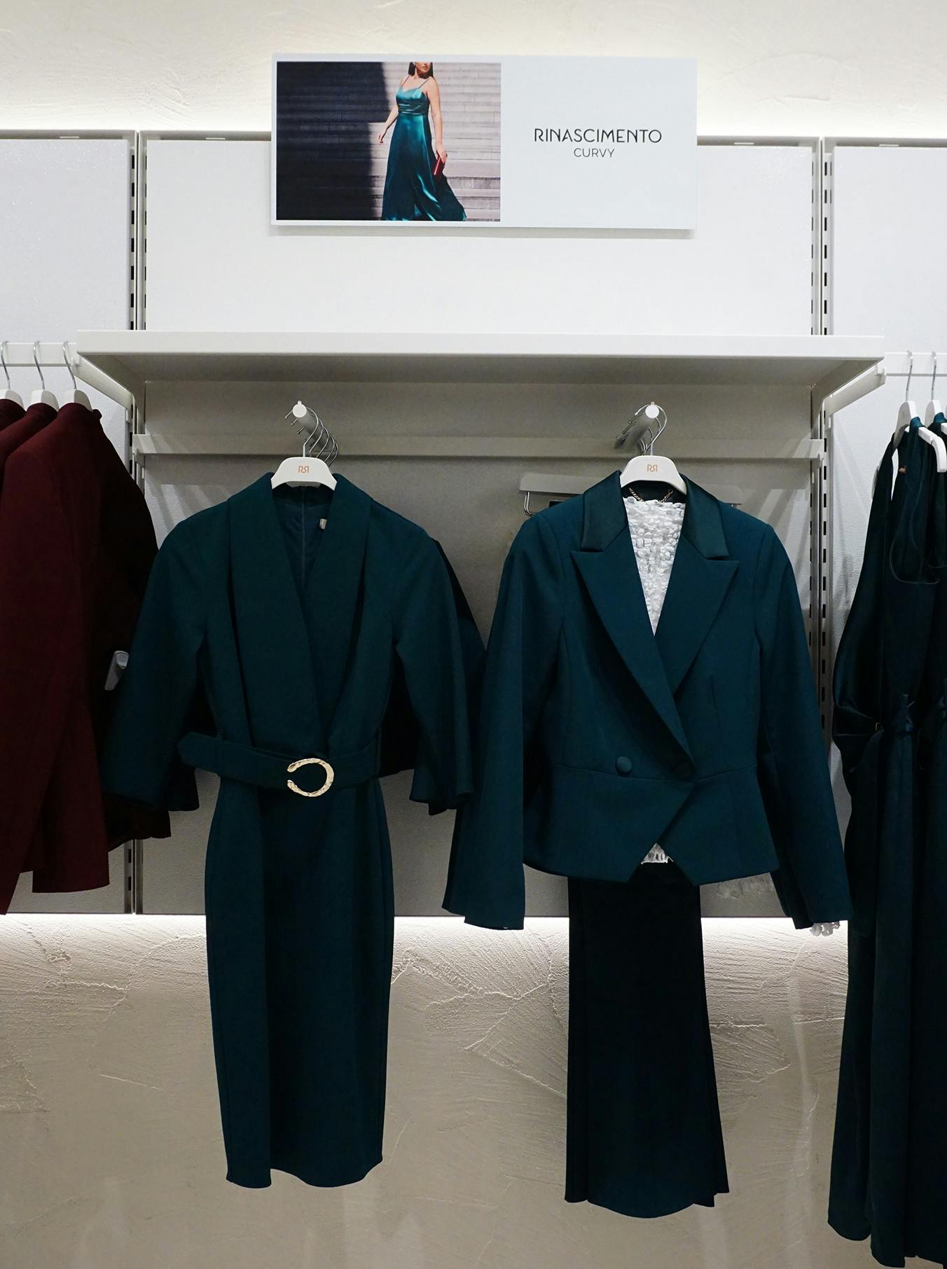 Dark teal formal outfits on hangers against a white backdrop with a "Rinascimento Curvy" sign and an elegant dress photo. Sophisticated display.