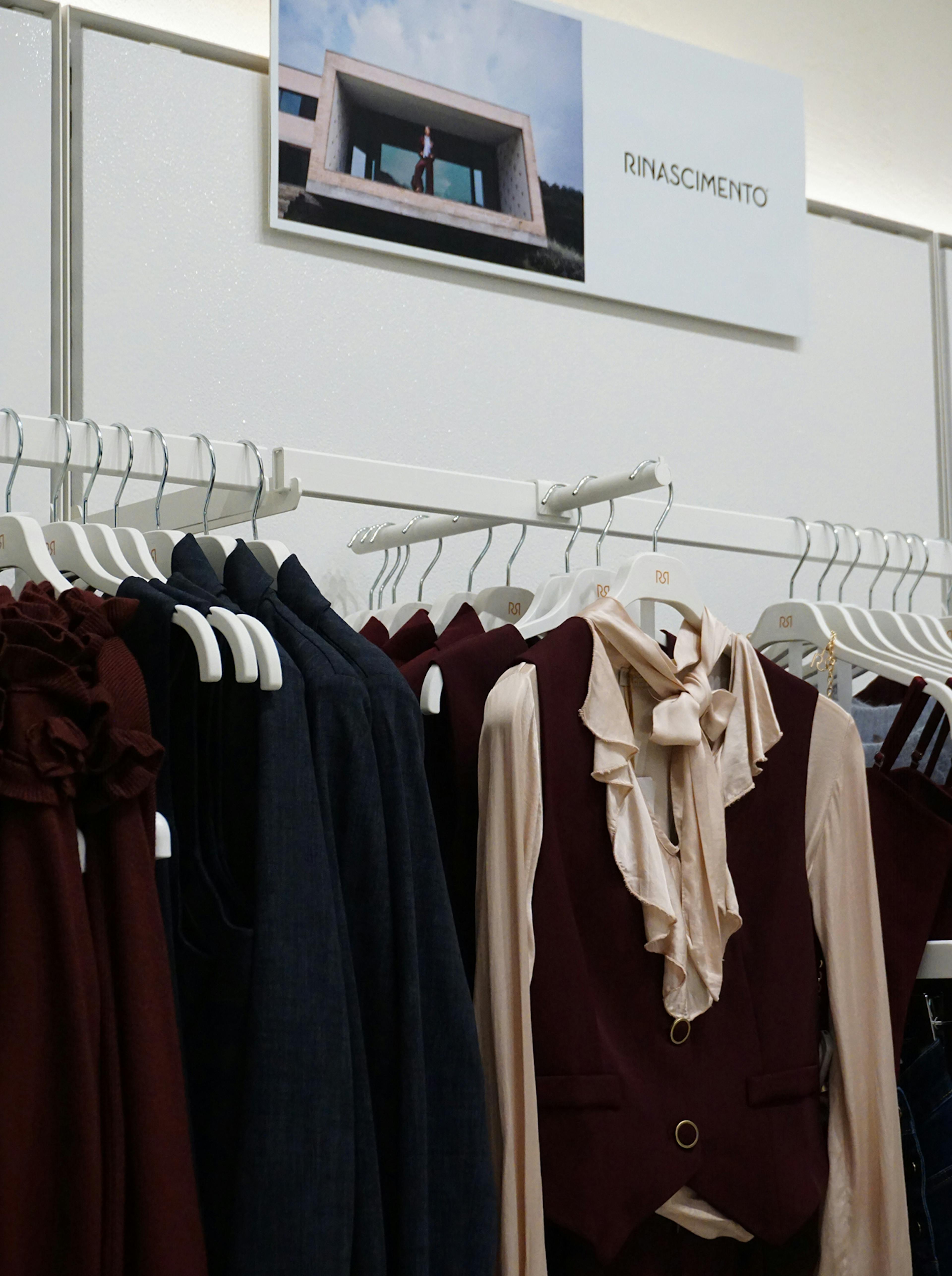 Clothing rack with elegant burgundy and navy garments, featuring ruffled blouses and vests. A Rinascimento sign is displayed above.