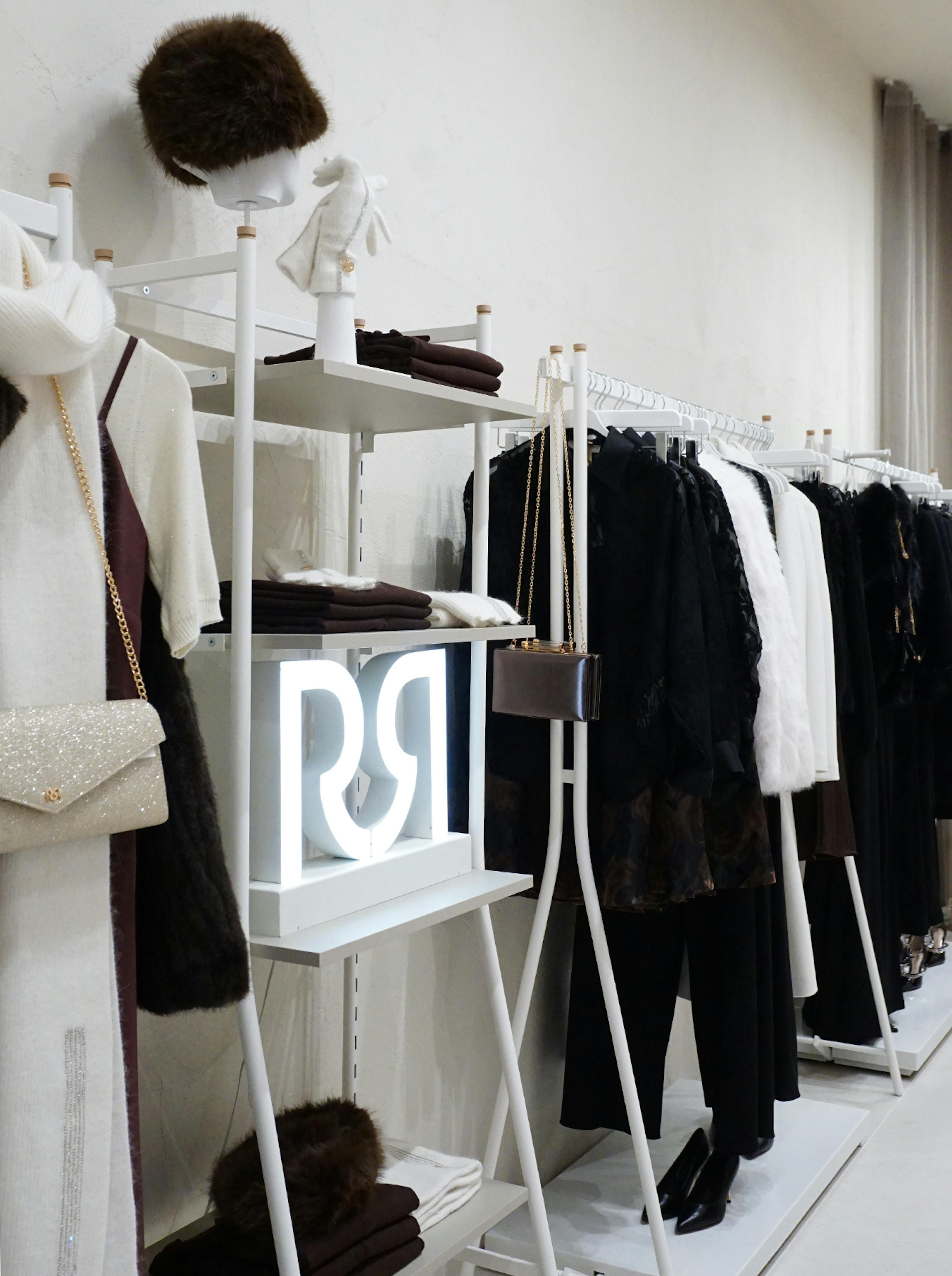 A chic boutique display features elegant winter clothing, including a white dress, coats, and accessories. A glowing sign adds a modern touch.