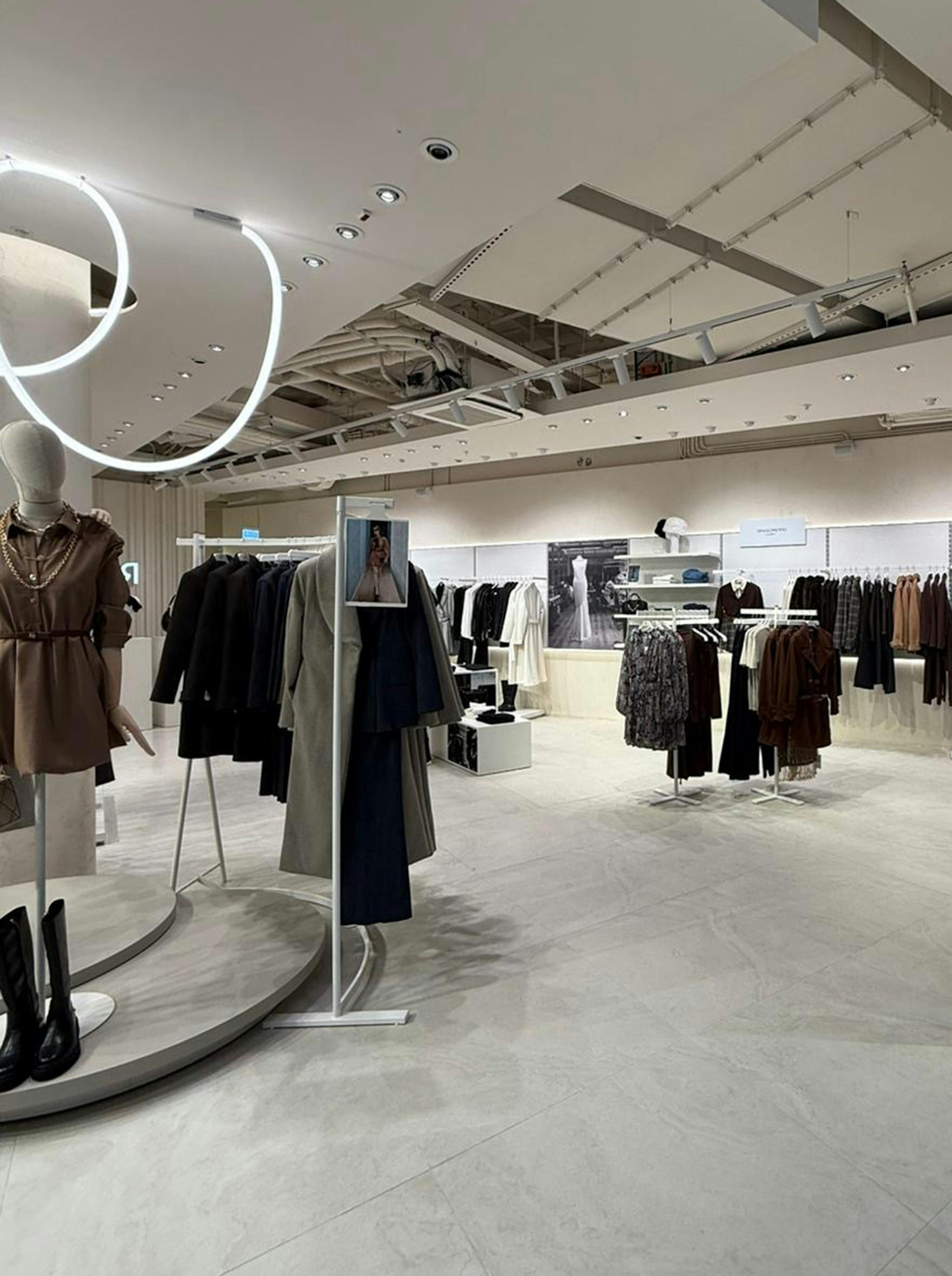 Modern clothing store interior with elegant displays of neutral-toned garments. A mannequin stands prominently, with circular ceiling lights enhancing the chic ambiance.