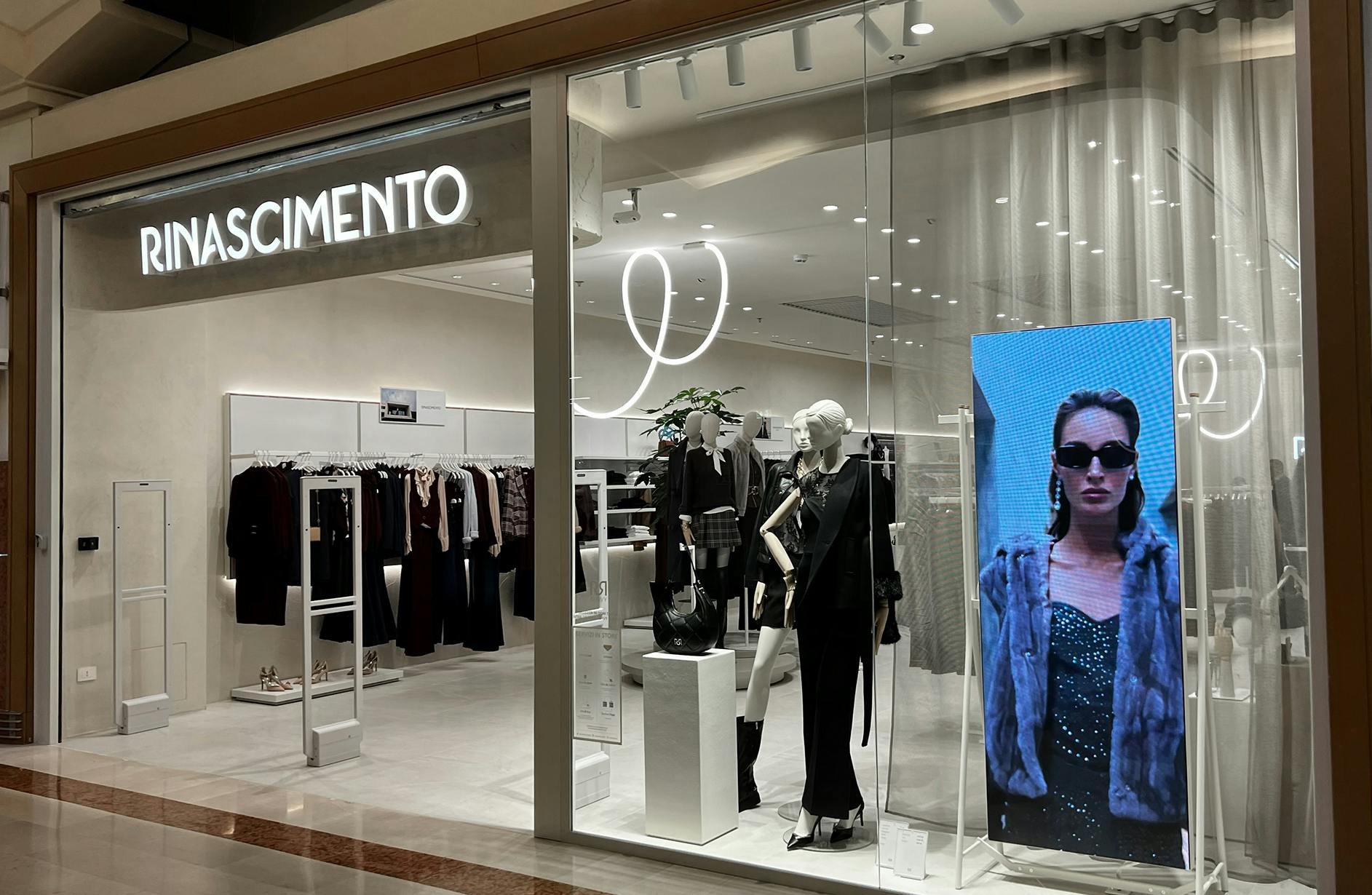 Storefront of RINASCIMENTO with mannequins in chic outfits and a digital display showing a model in sunglasses, conveying modern elegance.