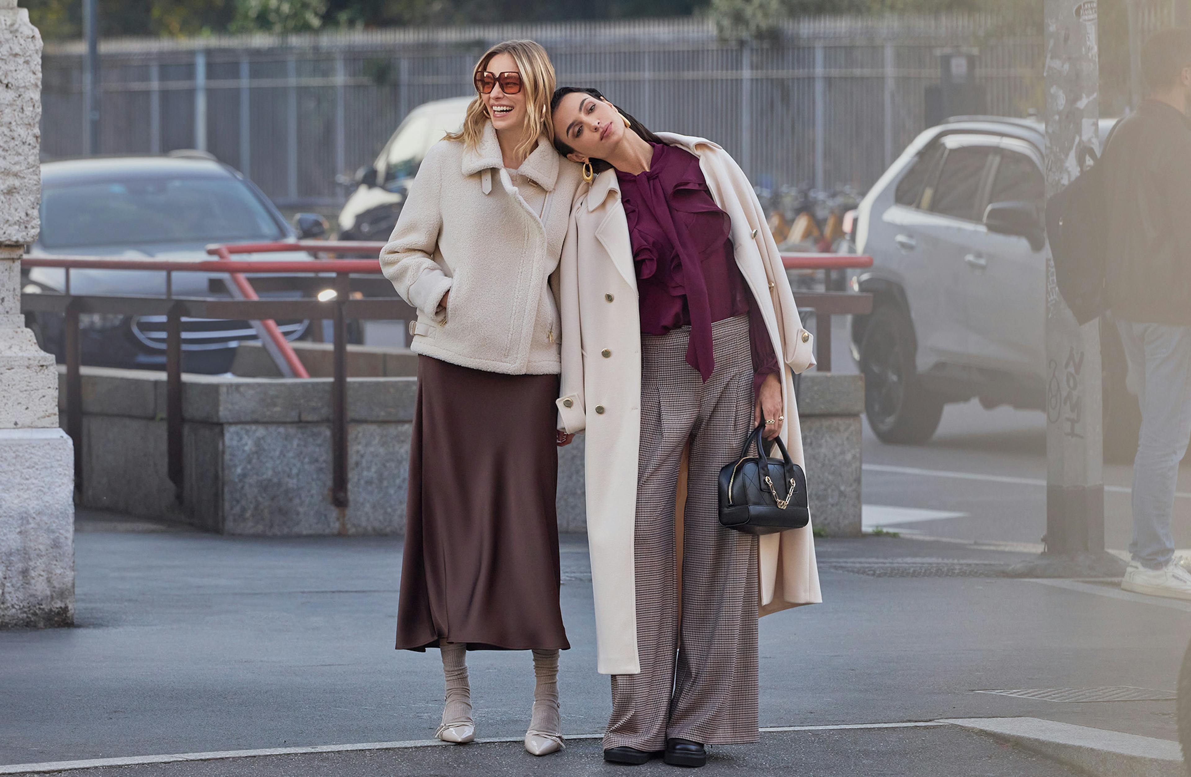 Two women in stylish winter outfits smile on a city street. One in a white coat and sunglasses, the other in a beige coat and burgundy blouse, exude joy and friendship.