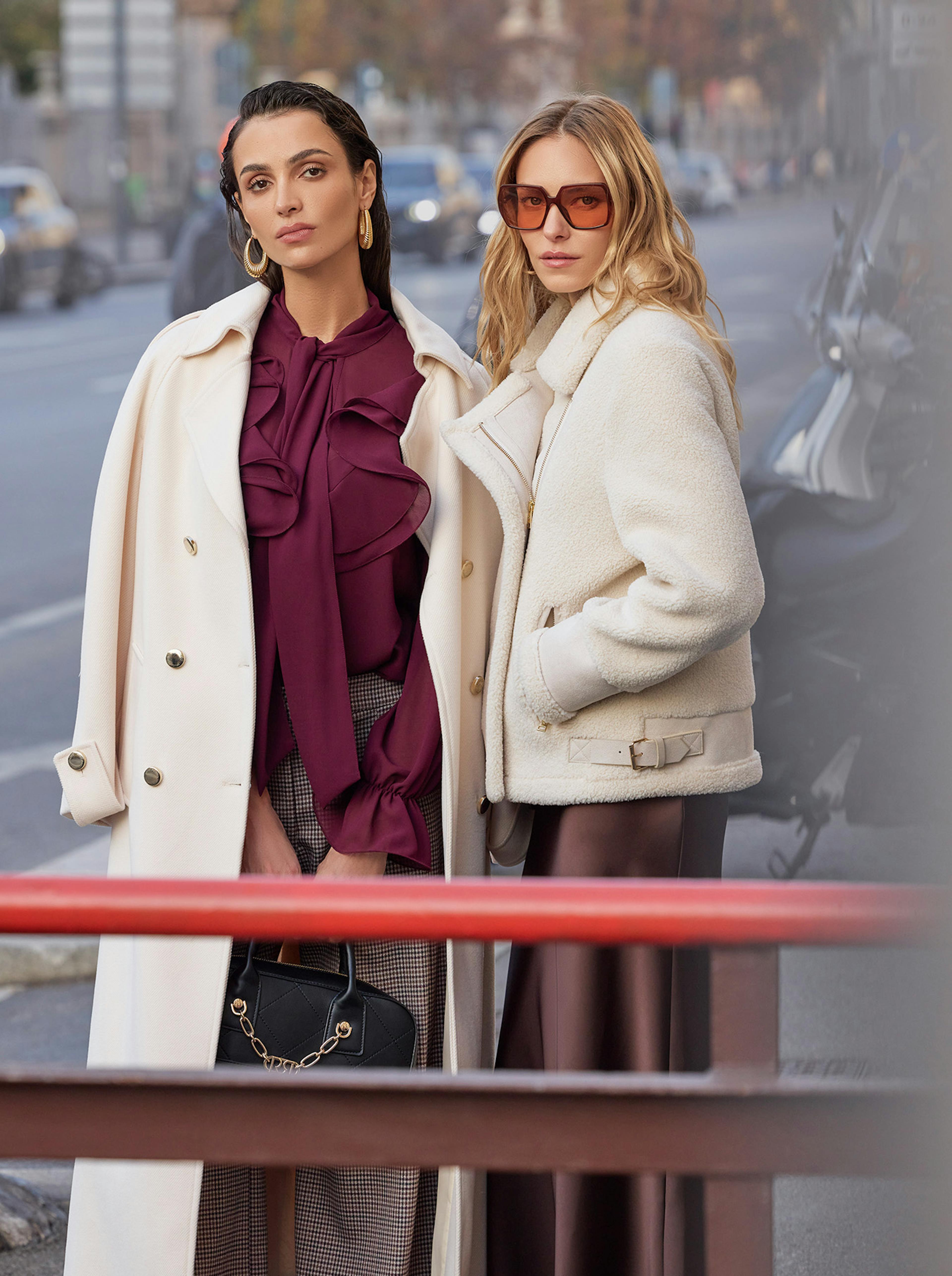 Two women in stylish winter outfits smile on a city street. One in a white coat and sunglasses, the other in a beige coat and burgundy blouse, exude joy and friendship.