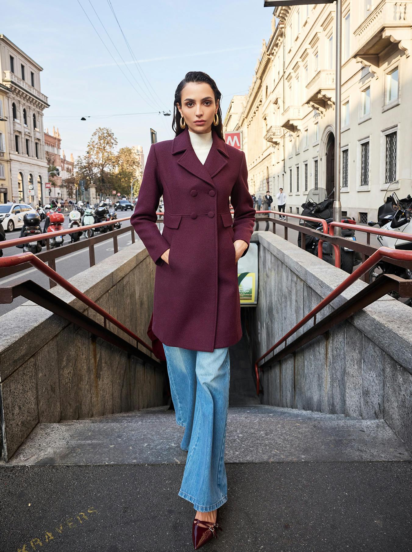 A person stands confidently at a city subway entrance, wearing a stylish burgundy coat, light blue jeans, and maroon heels. The street and buildings create an urban backdrop.
