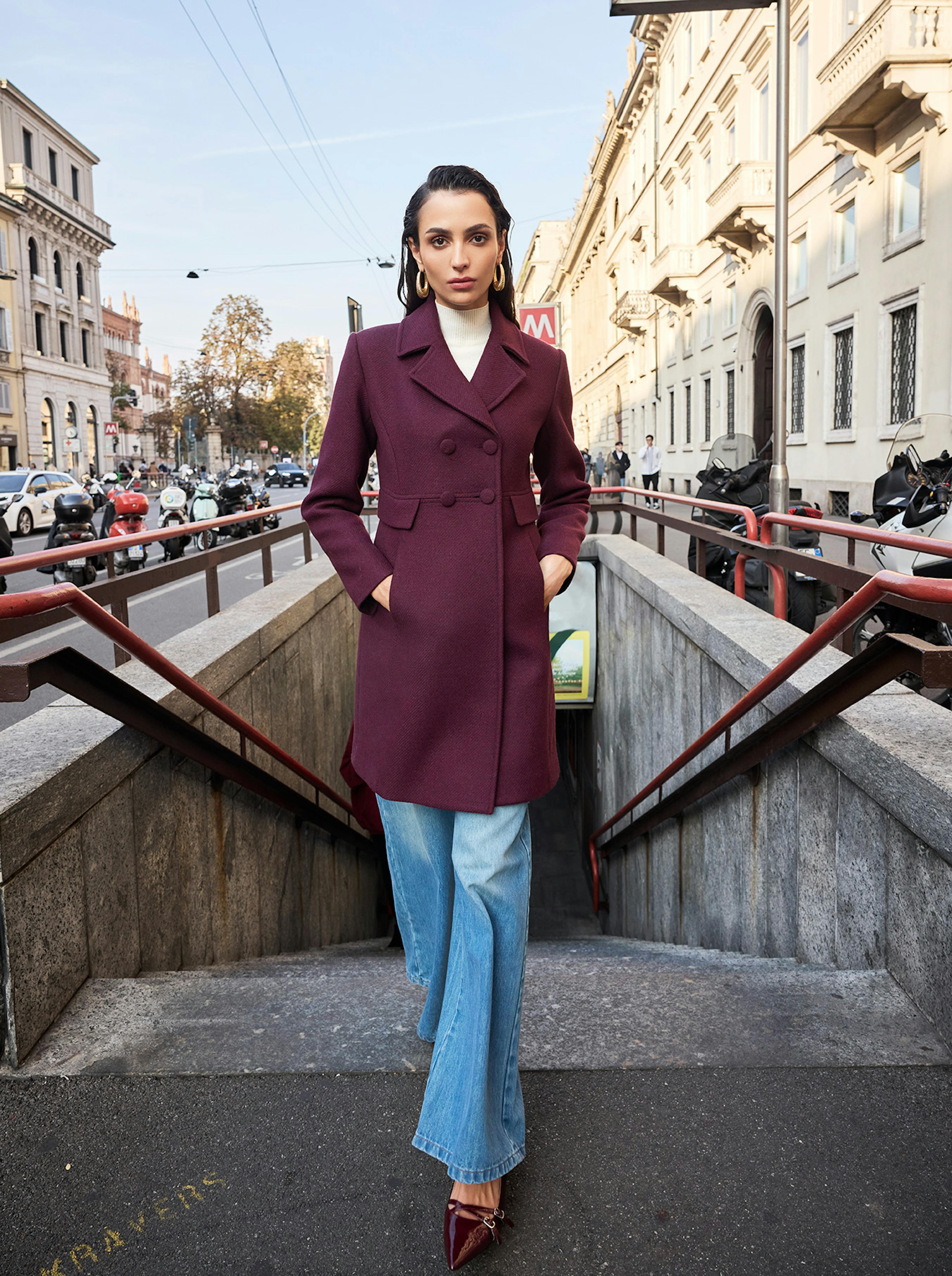 A person stands confidently at a city subway entrance, wearing a stylish burgundy coat, light blue jeans, and maroon heels. The street and buildings create an urban backdrop.