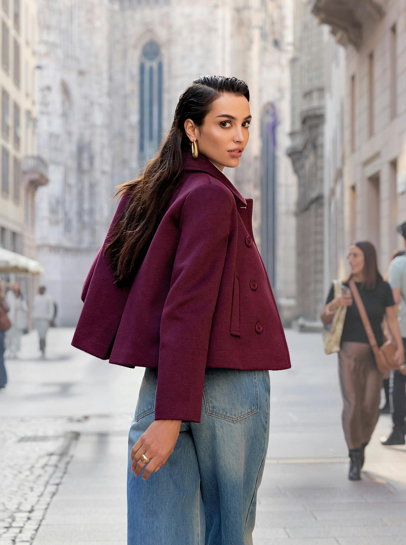 A woman in a stylish burgundy coat and jeans glances over her shoulder on a bustling street, with a historic cathedral blurred in the background.
