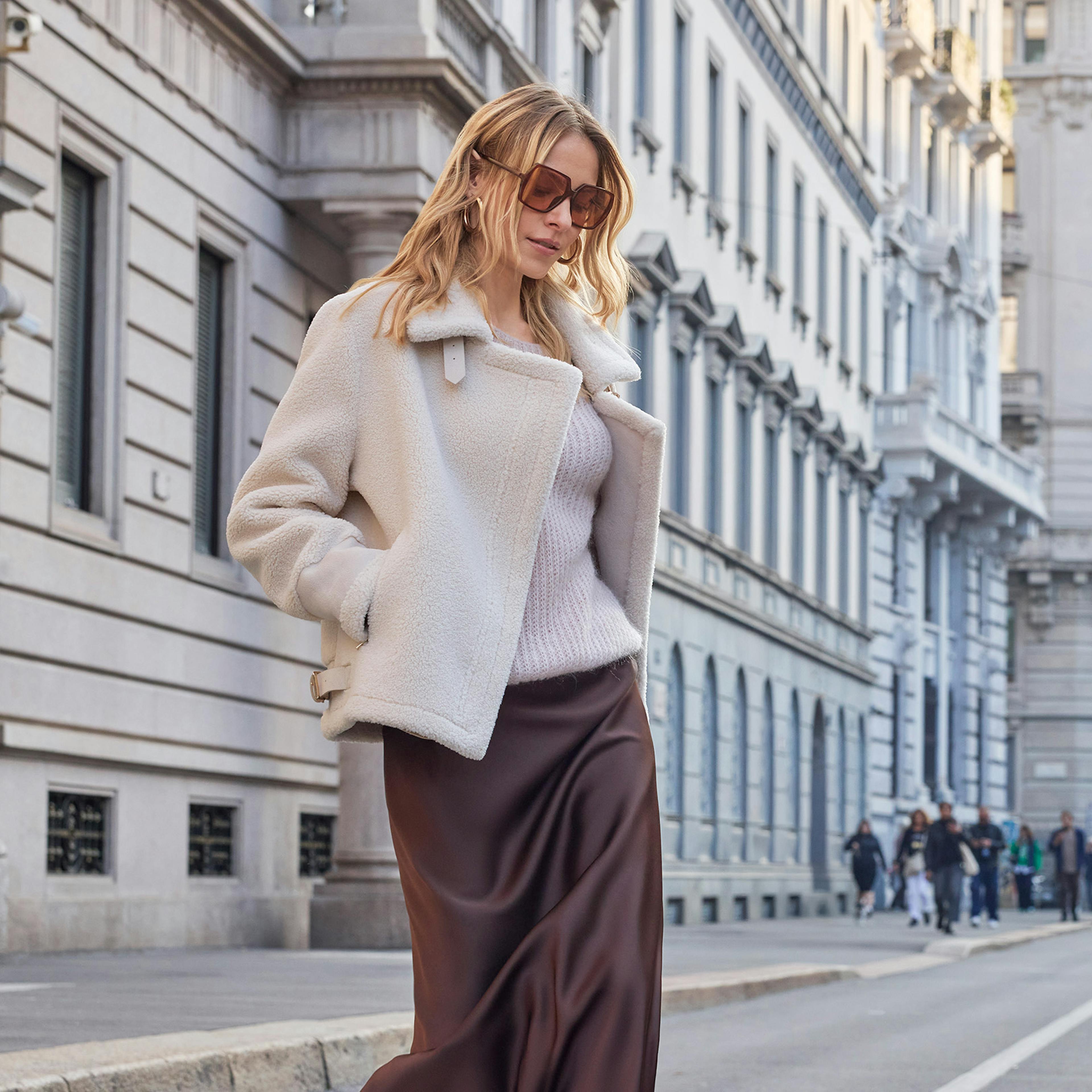 A woman in a chic outfit with a white coat and brown skirt walks confidently down a city street lined with historic buildings on a clear day.
