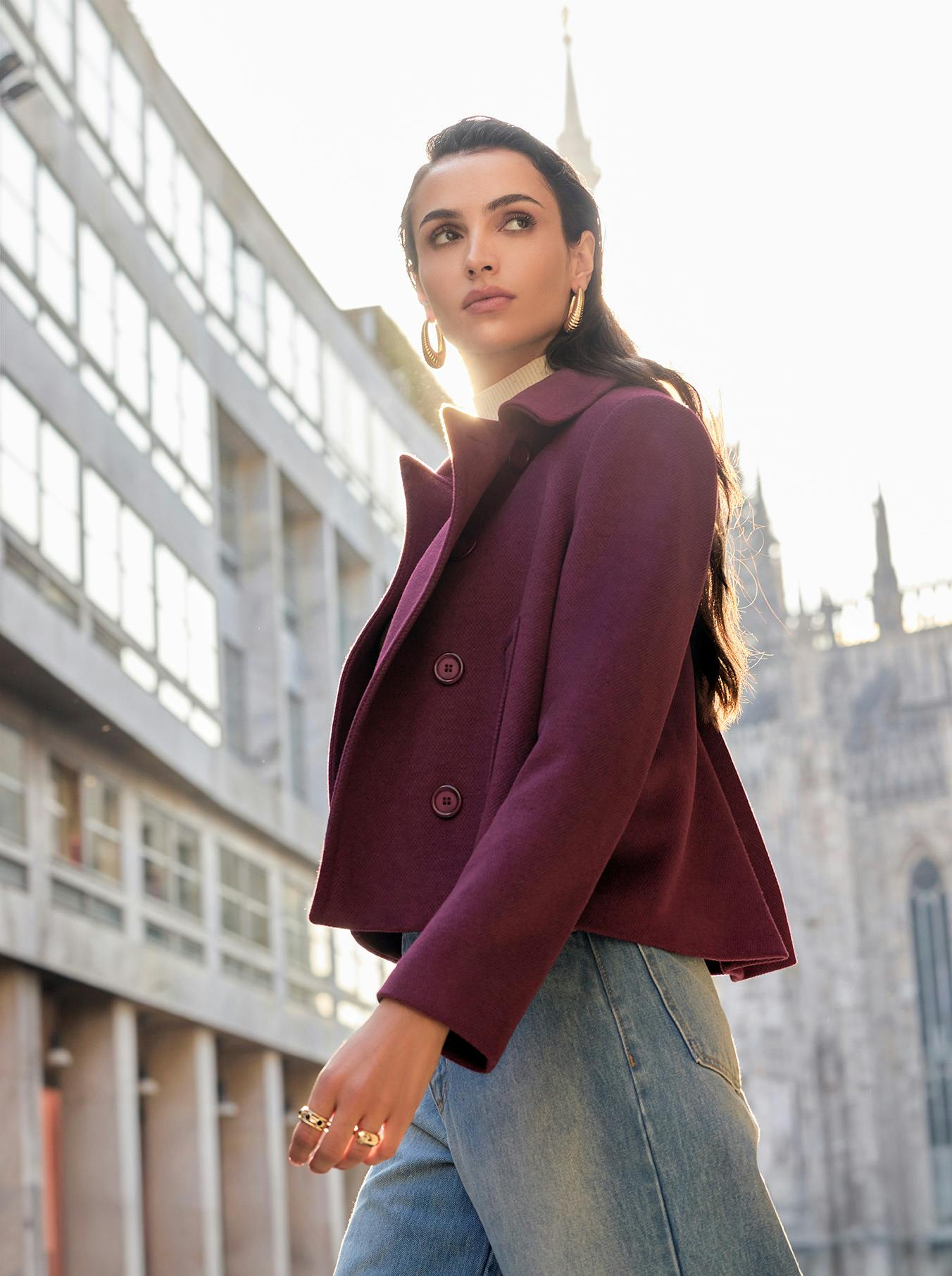 Woman in a burgundy coat and jeans stands confidently in an urban setting with modern buildings. Sunlight creates a warm, stylish ambiance.
