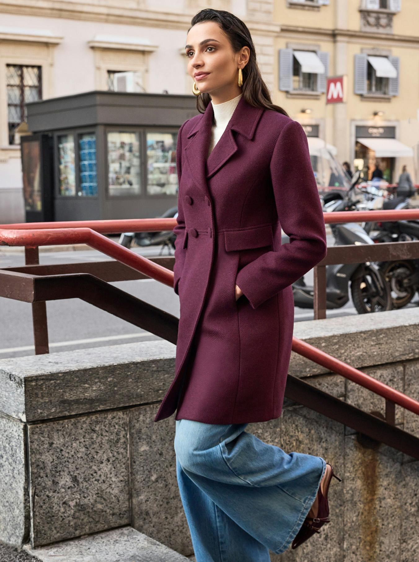 A woman in a burgundy coat and wide-leg jeans stands on city steps, looking off to the side. The urban background features blurred buildings.