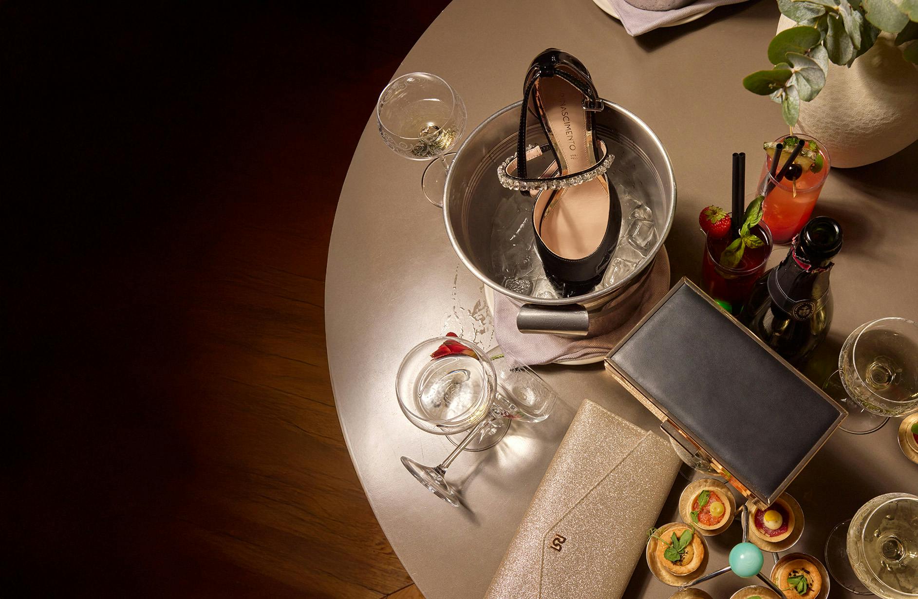 A high-heeled shoe rests in an ice bucket on a table with cocktails, appetizers, and a clutch, conveying a glamorous, party atmosphere.