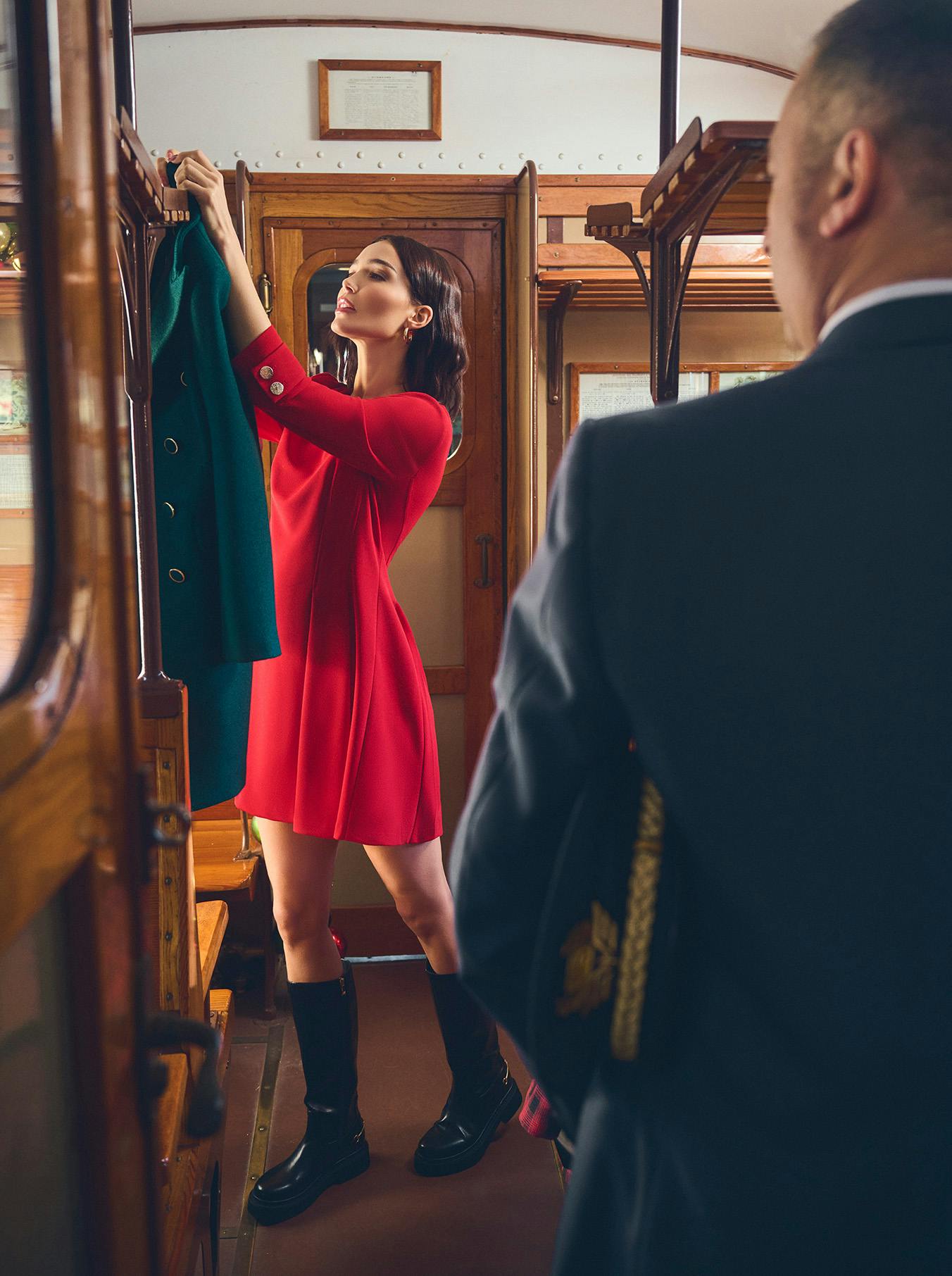 Woman in a red suit stands in a vintage train aisle, holding a handbag. Another woman, in plaid, sits nearby. Luggage and gift boxes are above. Elegant and nostalgic.