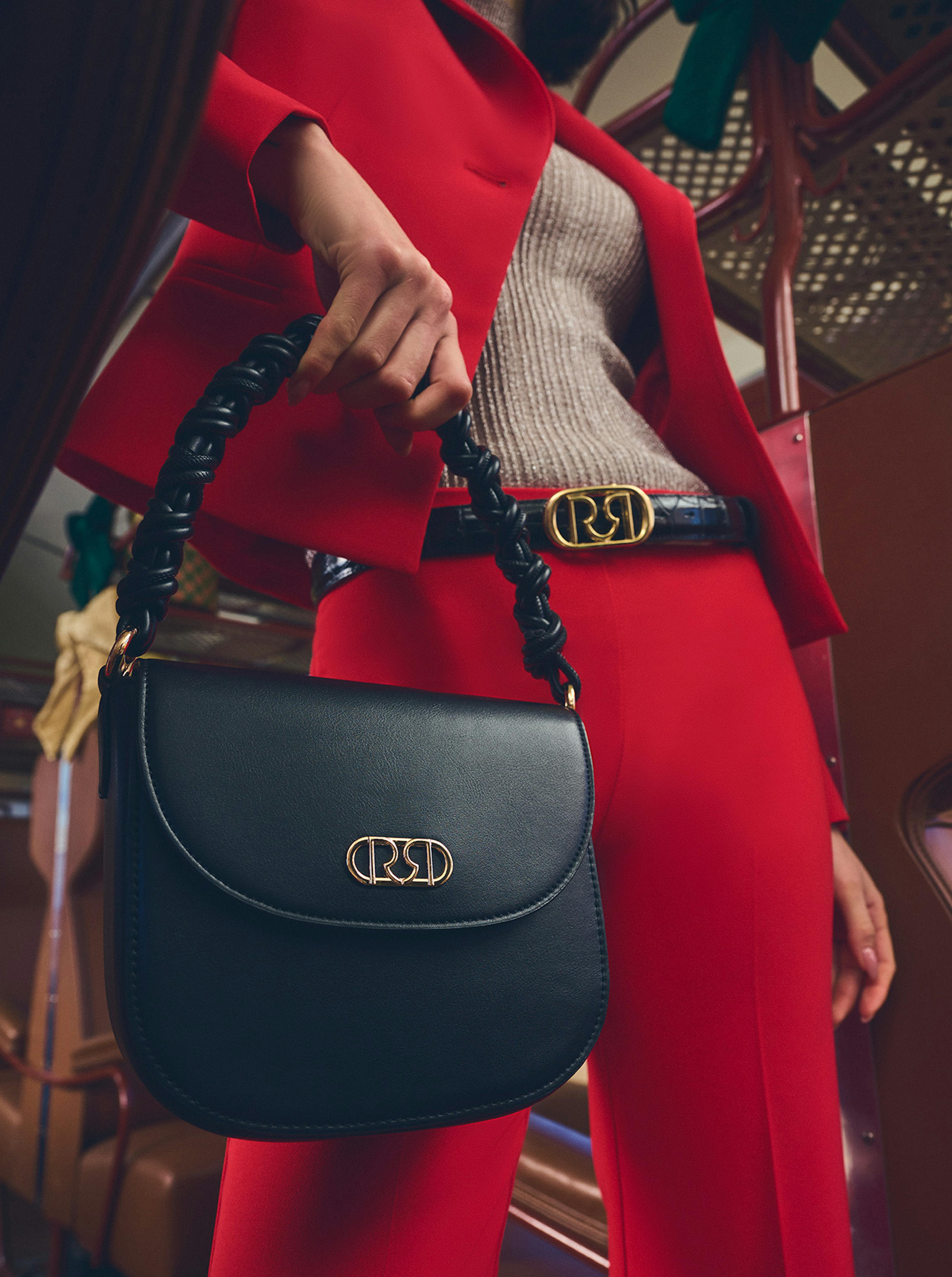 Woman in a red suit stands in a vintage train aisle, holding a handbag. Another woman, in plaid, sits nearby. Luggage and gift boxes are above. Elegant and nostalgic.
