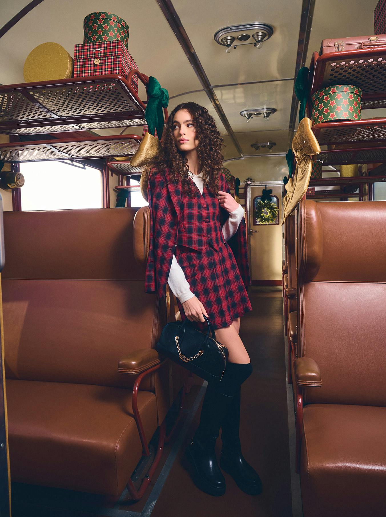 Young woman in a red plaid outfit stands confidently in a vintage train carriage with brown seats, surrounded by festive decorations and gifts.