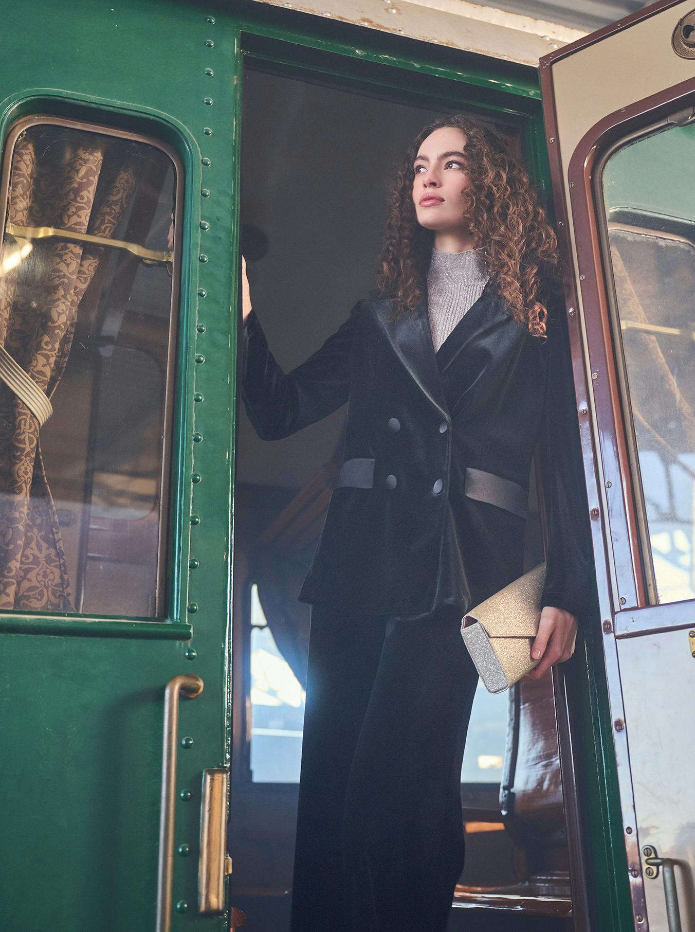 A woman in a black velvet outfit stands gracefully at the open door of a vintage green train carriage, holding a gold clutch. The scene is shrouded in mist, conveying a sense of elegance and nostalgia.
