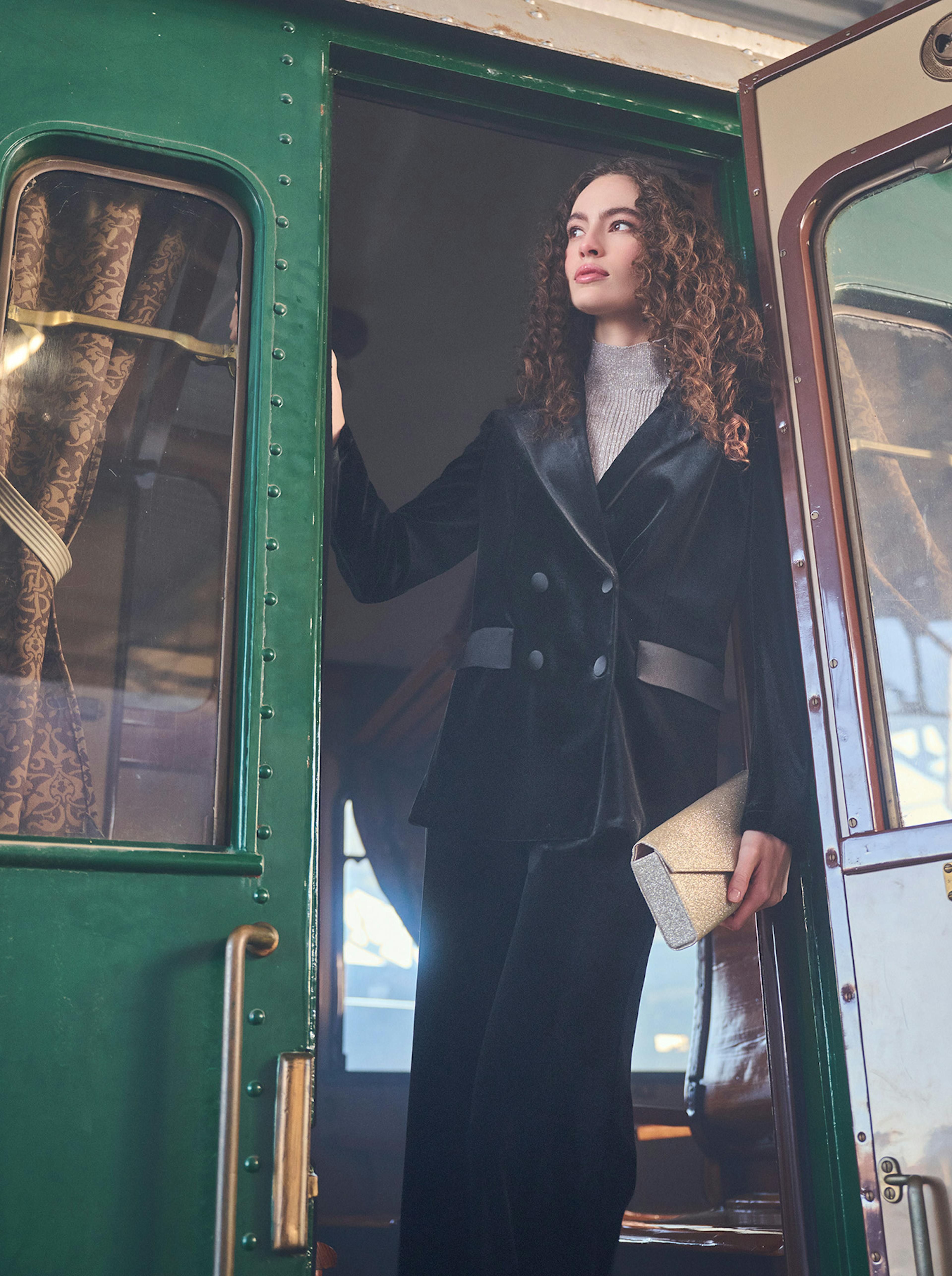 A woman in a black velvet outfit stands gracefully at the open door of a vintage green train carriage, holding a gold clutch. The scene is shrouded in mist, conveying a sense of elegance and nostalgia.