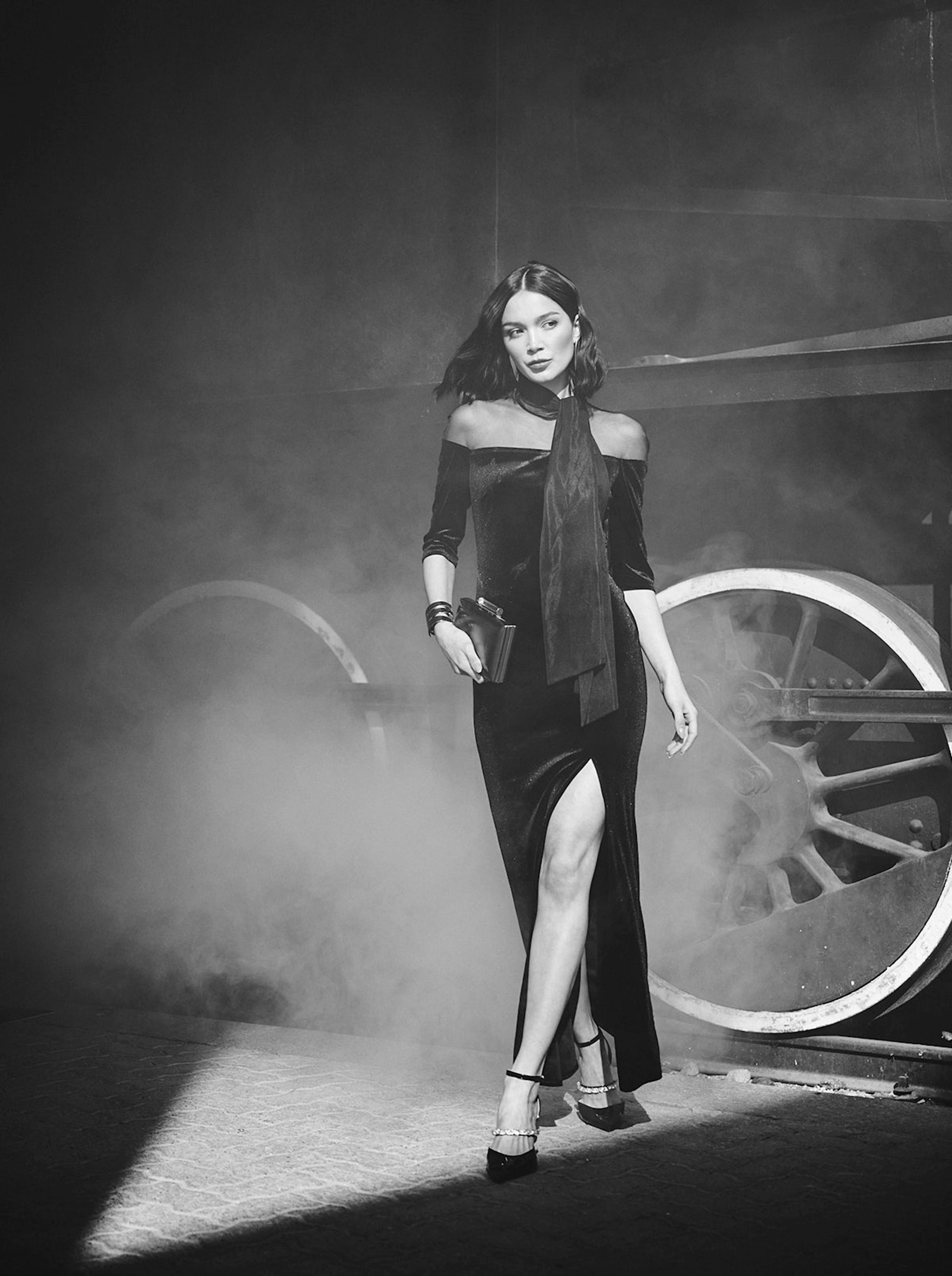 A woman in a sleek, black dress with a thigh-high slit walks confidently through a misty, industrial setting with large wheels, exuding elegance and strength.