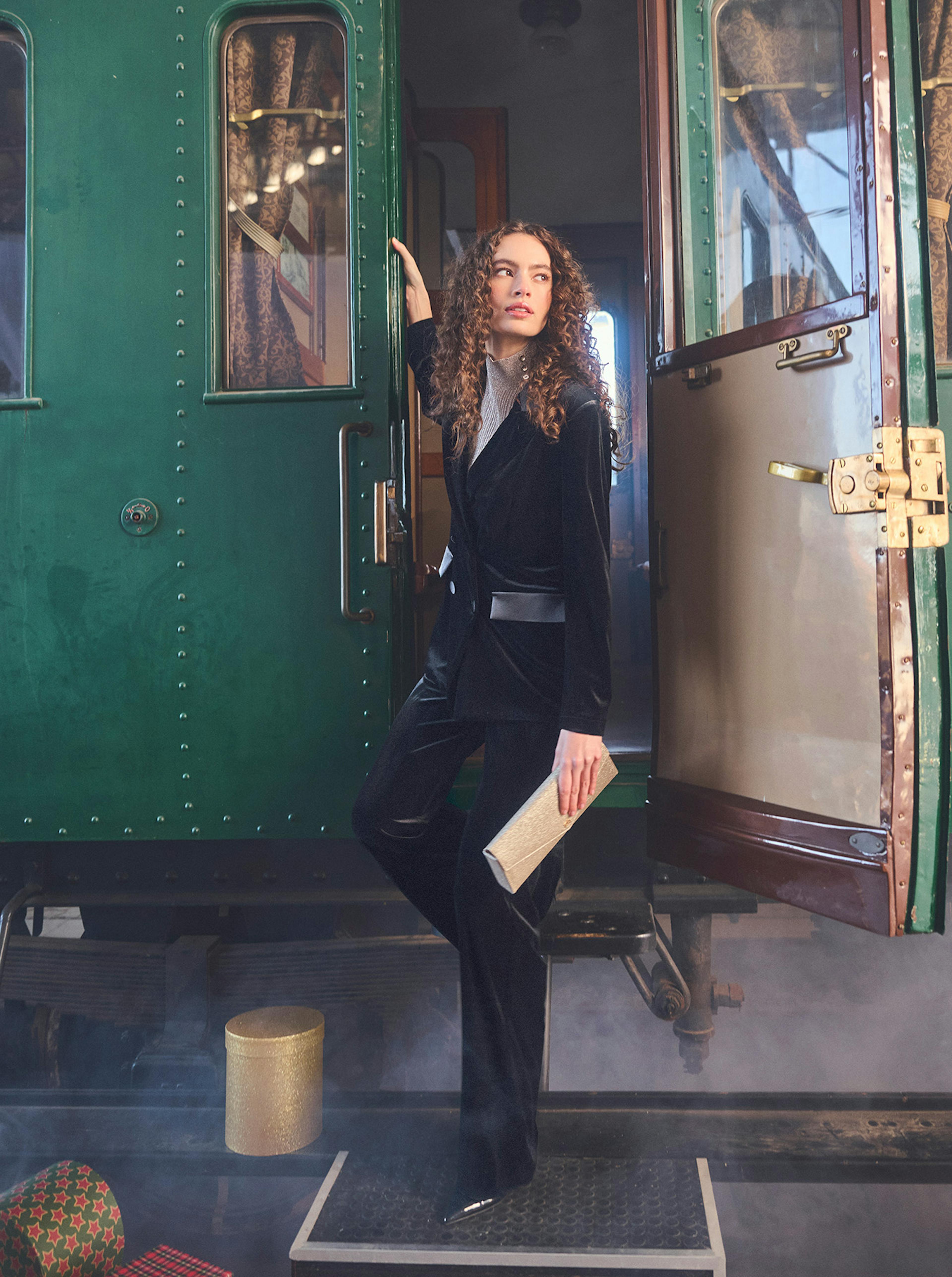 A woman in a black velvet outfit stands gracefully at the open door of a vintage green train carriage, holding a gold clutch. The scene is shrouded in mist, conveying a sense of elegance and nostalgia.