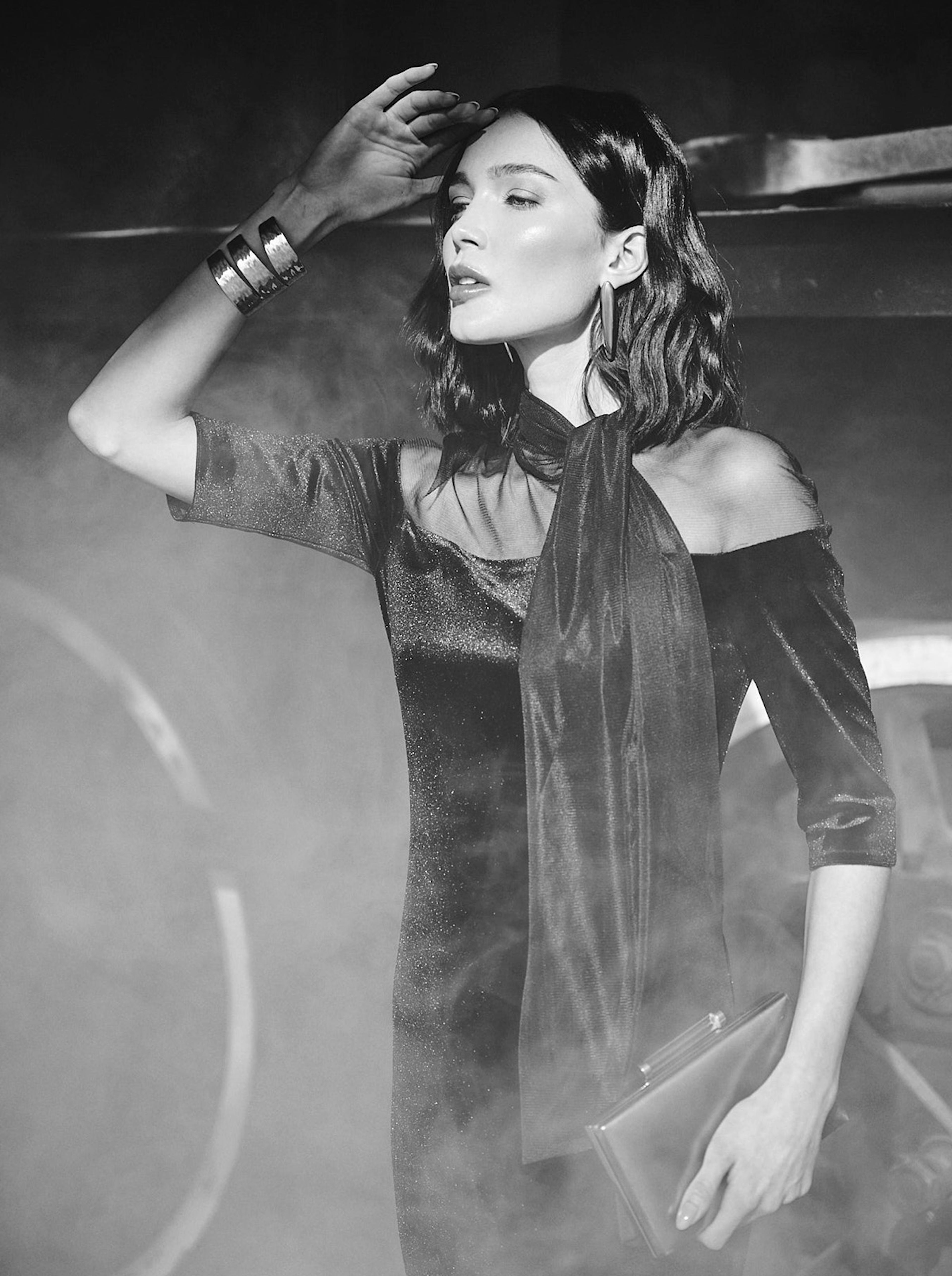 Stylish woman in an elegant, off-shoulder dress poses dramatically in smoky ambiance. She holds a clutch and wears bold jewelry, exuding sophistication.