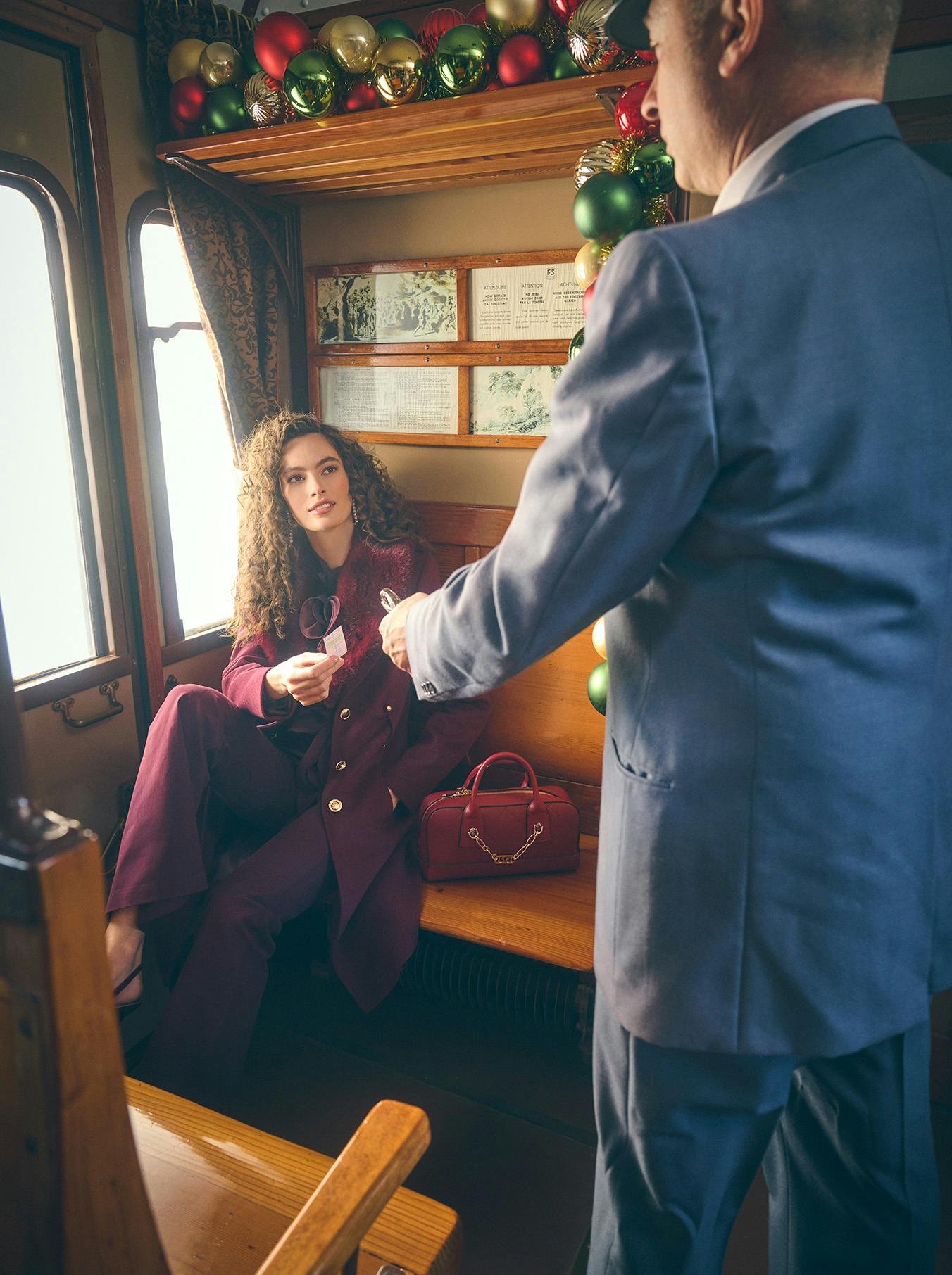 A woman in a burgundy suit sits on a vintage train seat, holding a ticket. A conductor in a blue suit stands nearby. Festive ornaments adorn the cabin.