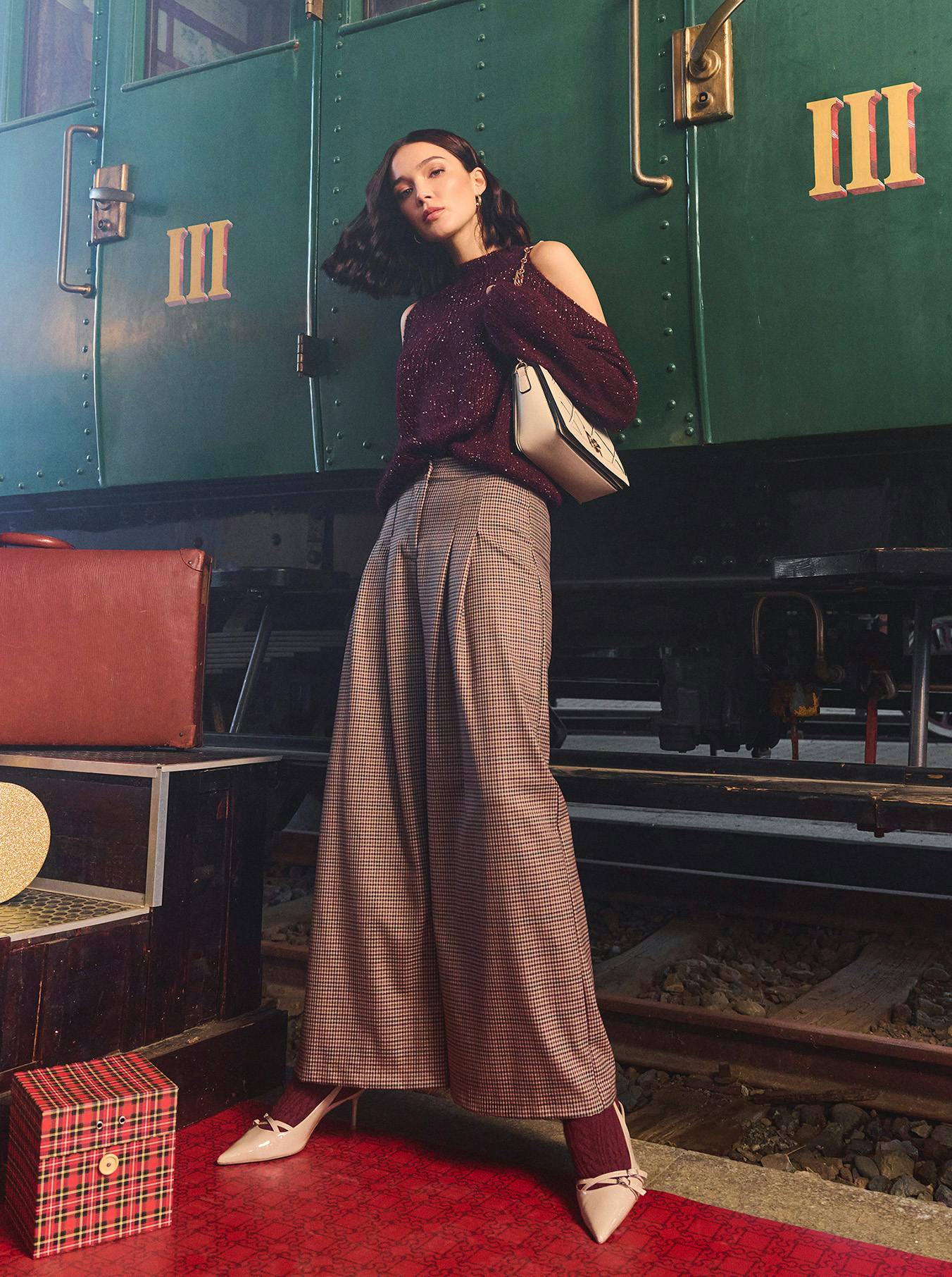 A woman stands confidently against a vintage train, wearing a burgundy top and plaid pants. Luggage is nearby, creating a retro travel vibe.