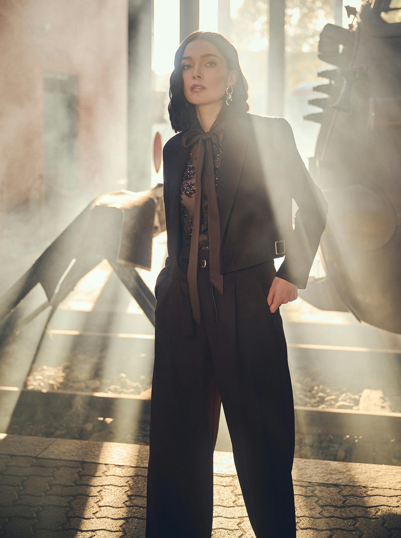 A woman stands confidently in a sunlit, smoky industrial setting, wearing an elegant black outfit with a sheer blouse. Sun rays highlight her poised expression.
