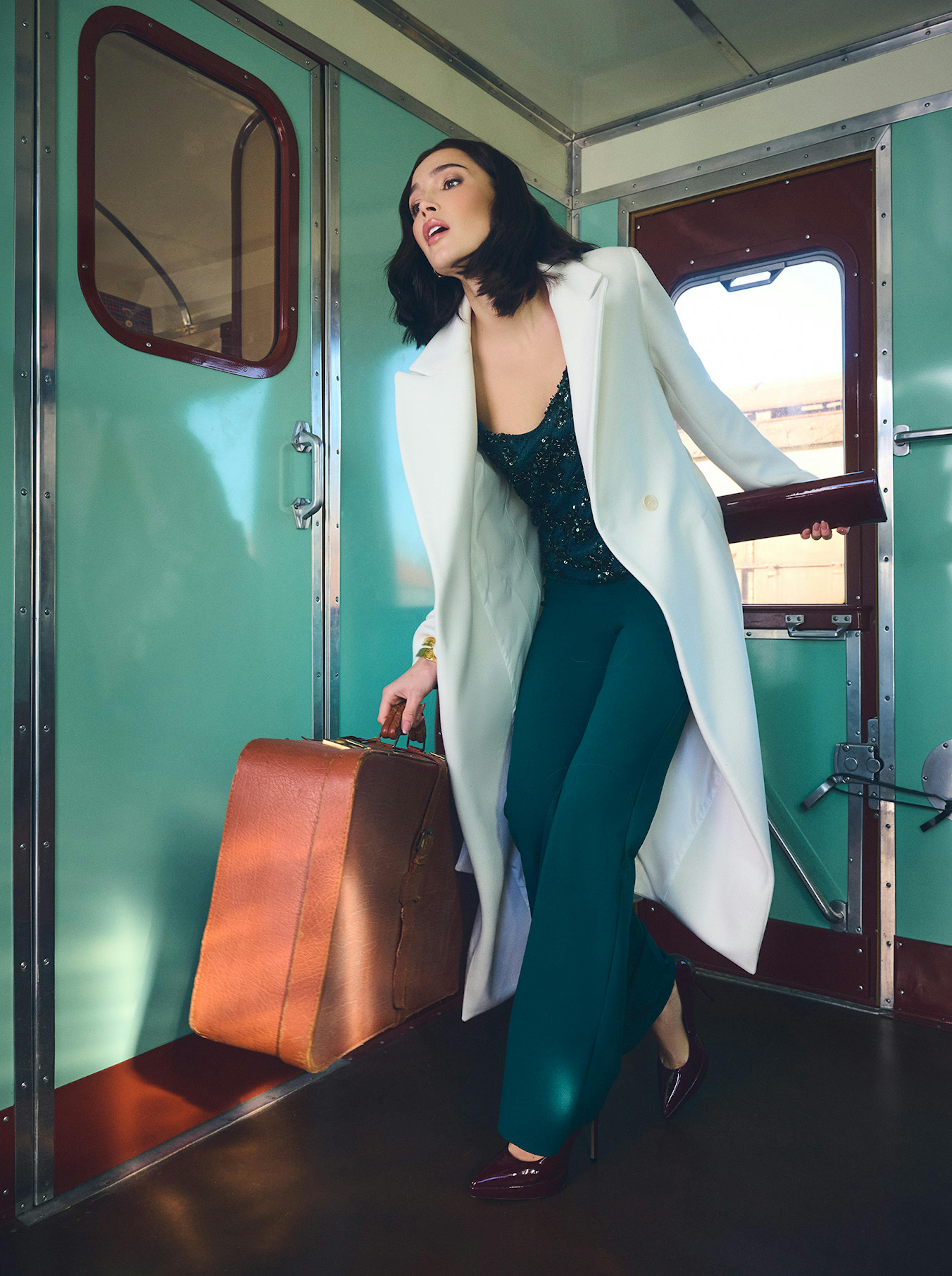 Woman with shoulder-length hair in a white coat and teal outfit, holding a brown suitcase and clutch in a vintage train, appears stylish and poised.