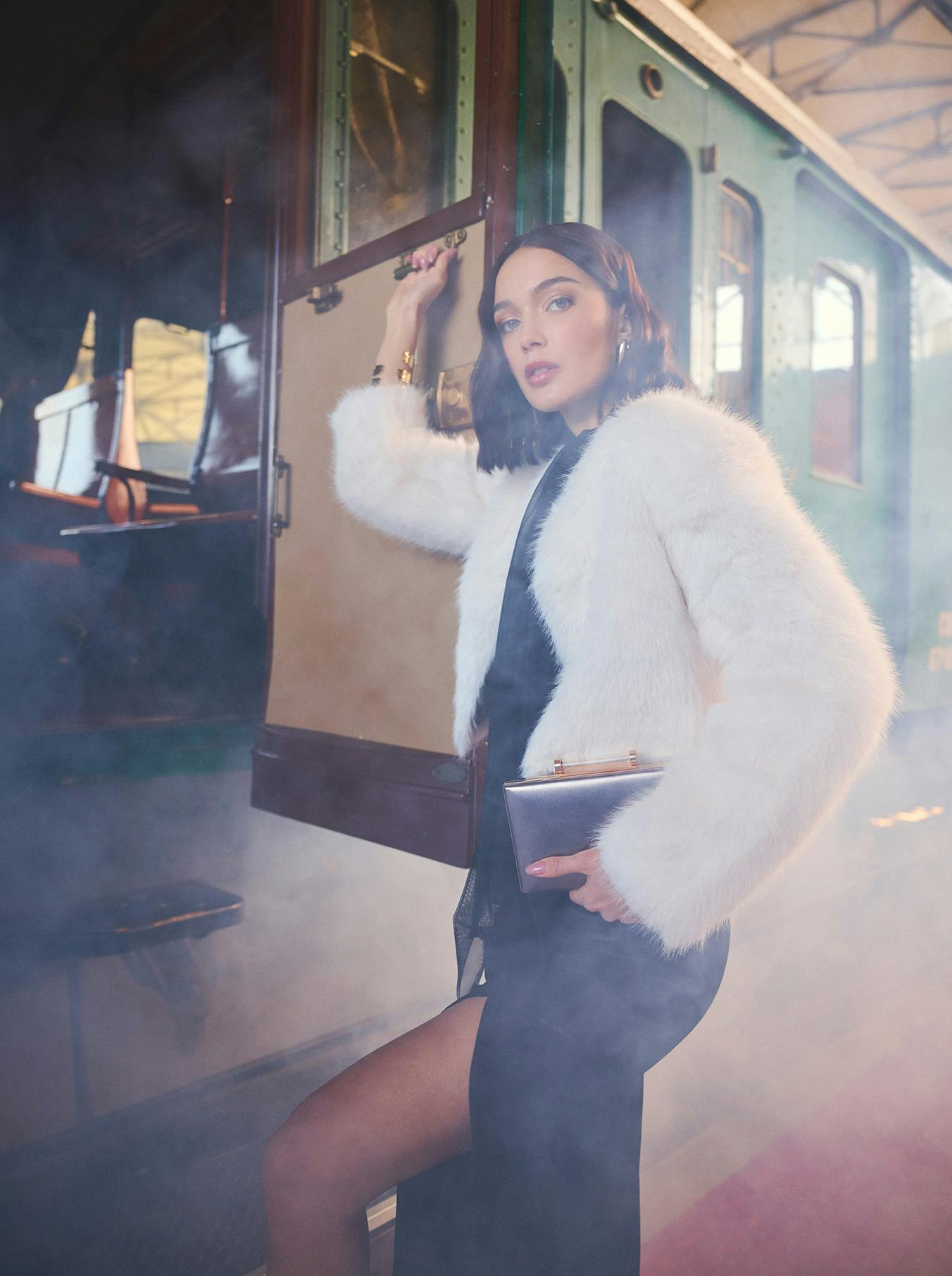 Woman in elegant black dress and white fur coat stands beside vintage train amid atmospheric mist, exuding a sense of glamour and nostalgia.