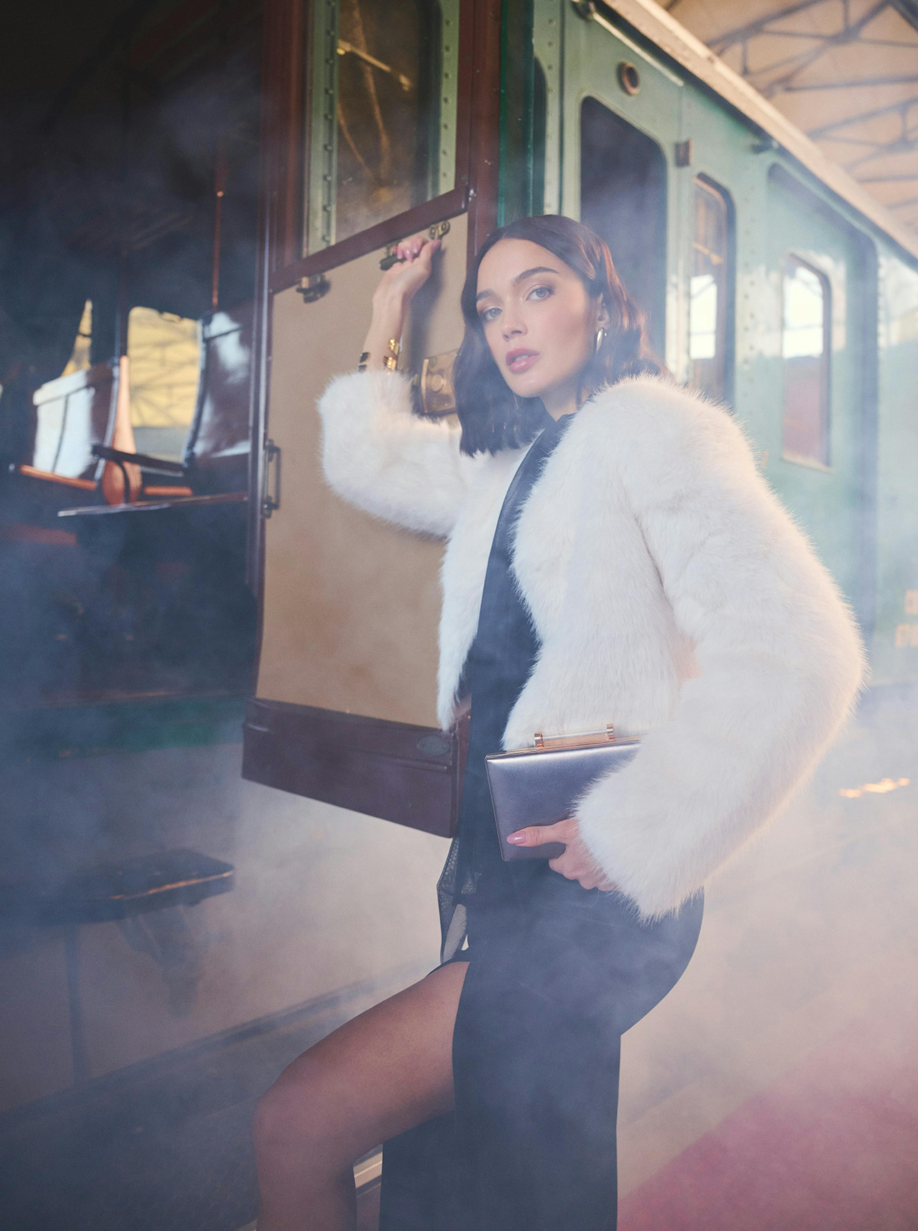 Woman in elegant black dress and white fur coat stands beside vintage train amid atmospheric mist, exuding a sense of glamour and nostalgia.