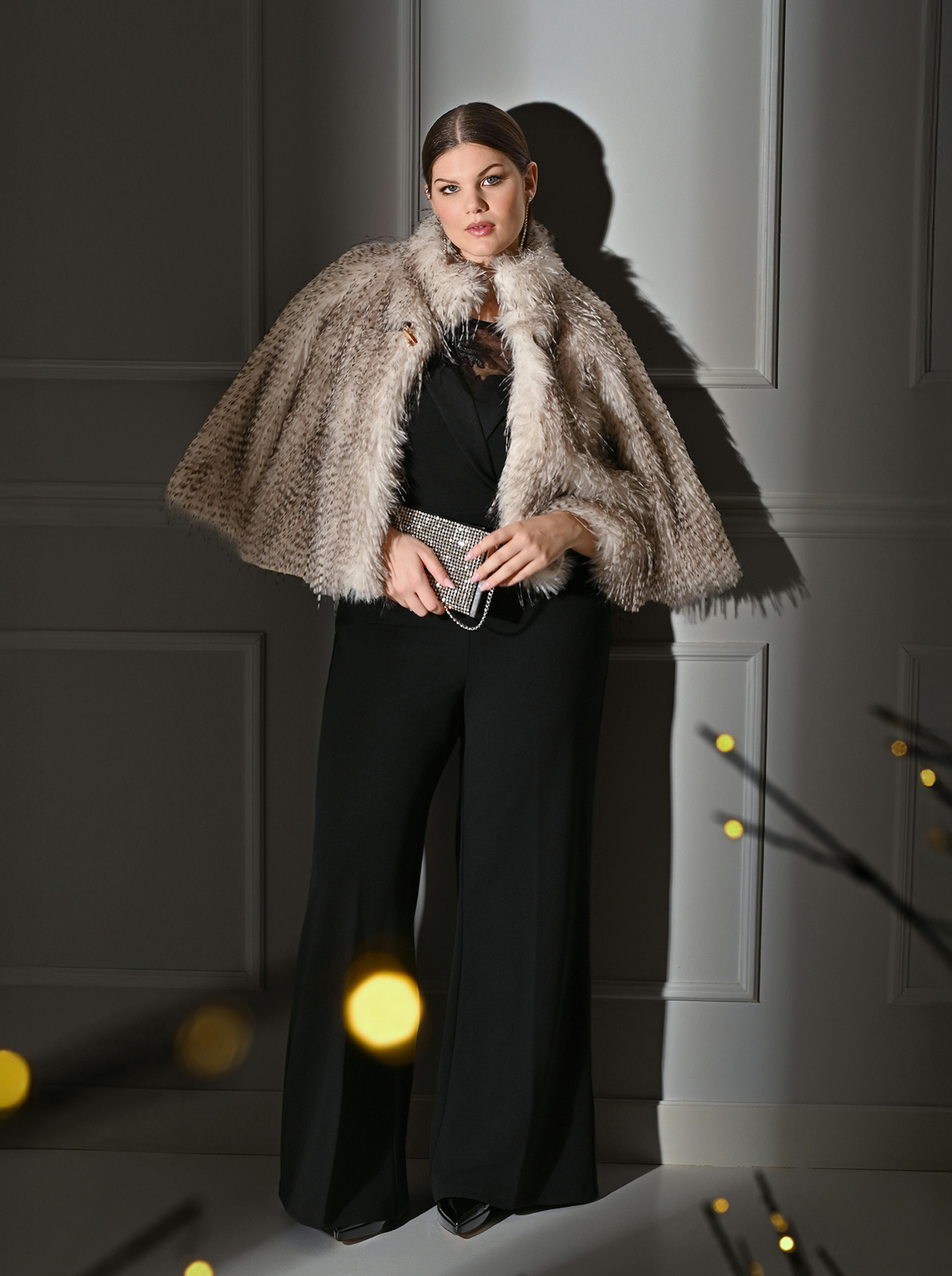 Woman in a black lace-detailed jumpsuit stands elegantly against a gray paneled wall. She holds a clutch, with warm bokeh lights in the foreground.