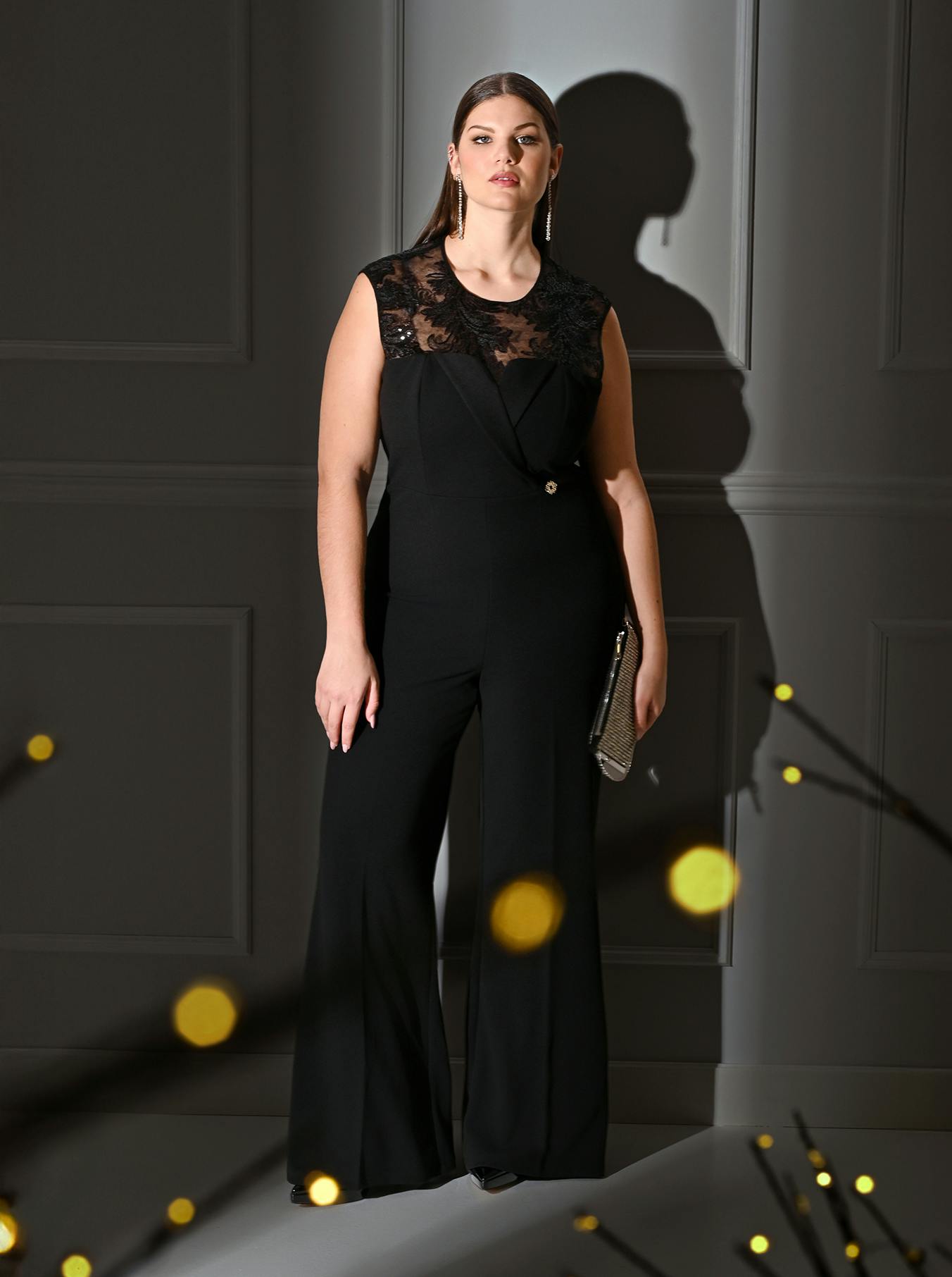 Woman in a black lace-detailed jumpsuit stands elegantly against a gray paneled wall. She holds a clutch, with warm bokeh lights in the foreground.
