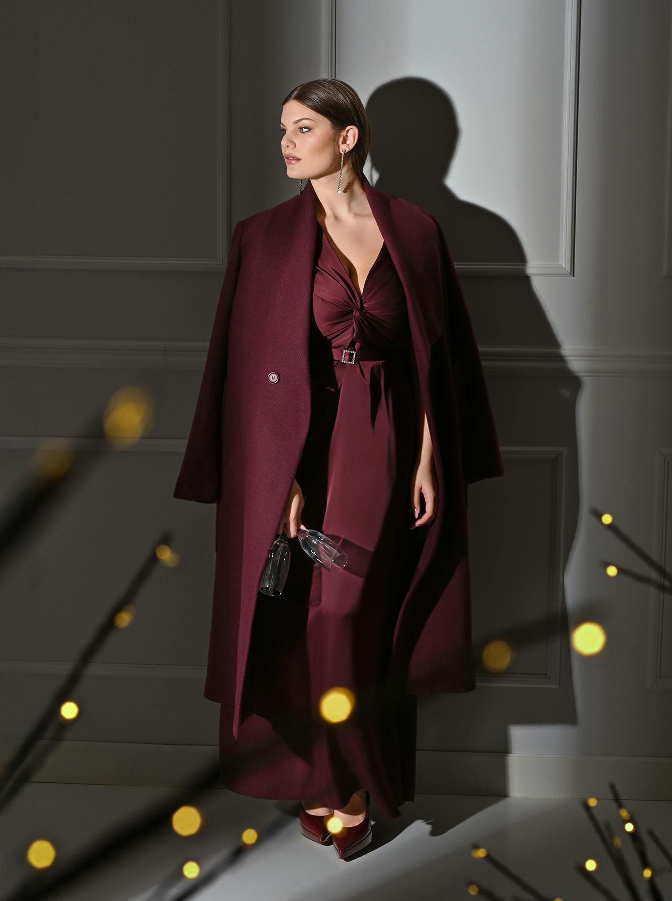Woman in a burgundy dress and coat stands against a paneled wall, holding a clutch. Ambient lighting with blurred yellow bokeh creates an elegant tone.