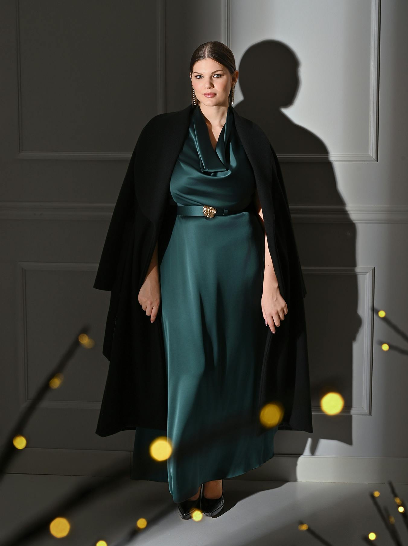 A woman in an elegant teal silk gown and black cape stands confidently against a gray wall. The setting is softly lit, with a serene and sophisticated atmosphere.