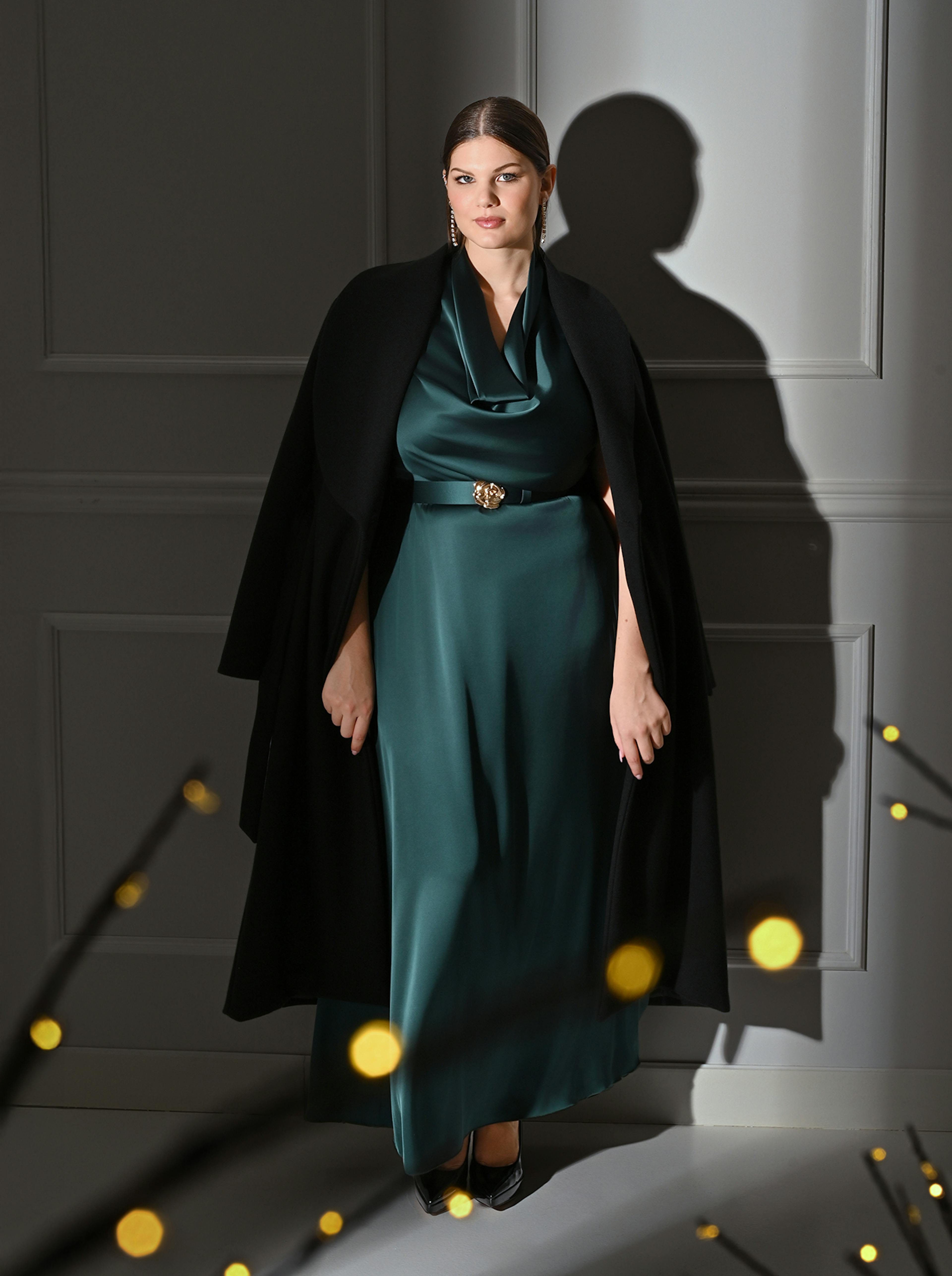 A woman in an elegant teal silk gown and black cape stands confidently against a gray wall. The setting is softly lit, with a serene and sophisticated atmosphere.