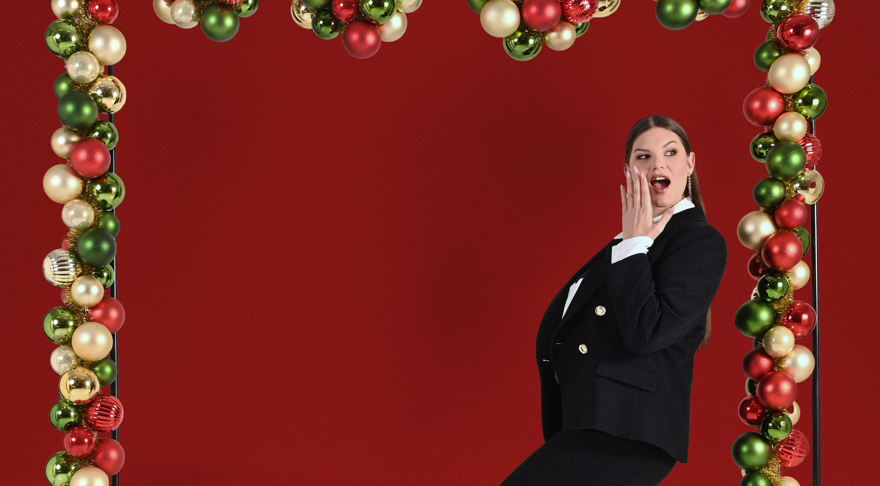 A woman in a black suit poses playfully under an arch decorated with red, green, and gold ornaments against a festive red background.