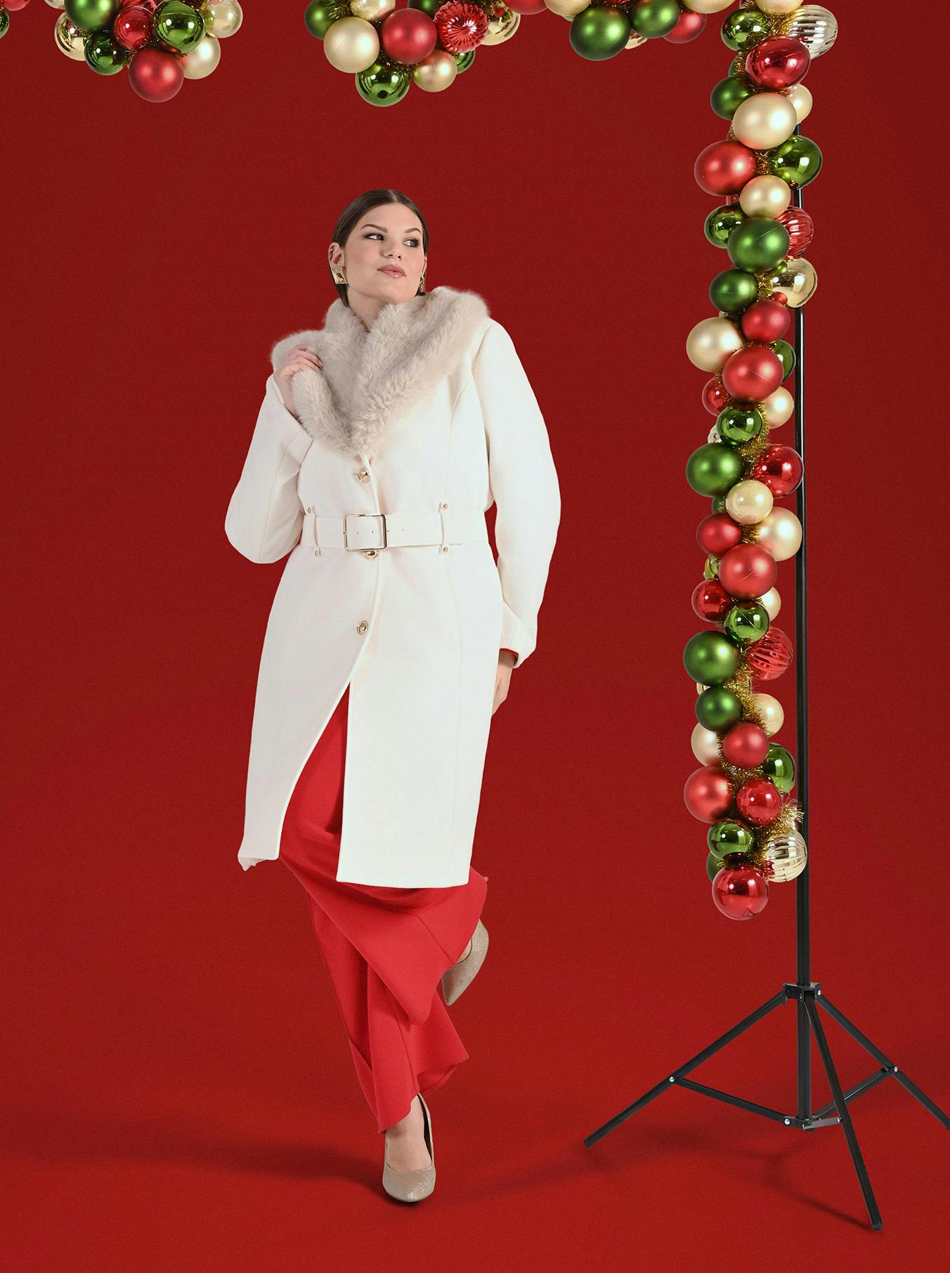 A woman in a white coat with a fur collar and red pants stands on a red background. She's next to a holiday arch made of red, green, and gold ornaments.
