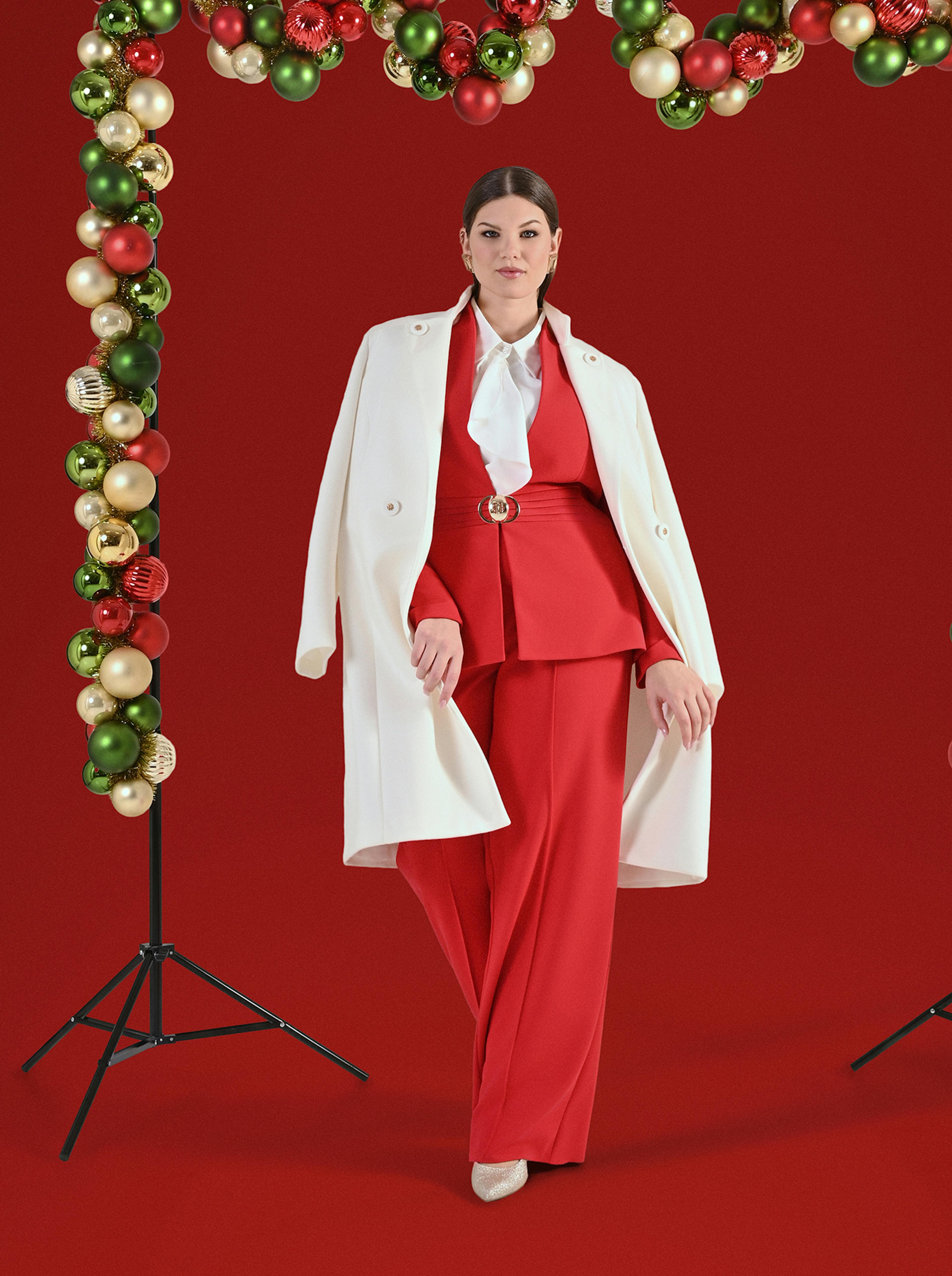 A woman in a striking red suit with a white blouse and coat stands confidently against a red background. Decorative green and red ornaments frame her.