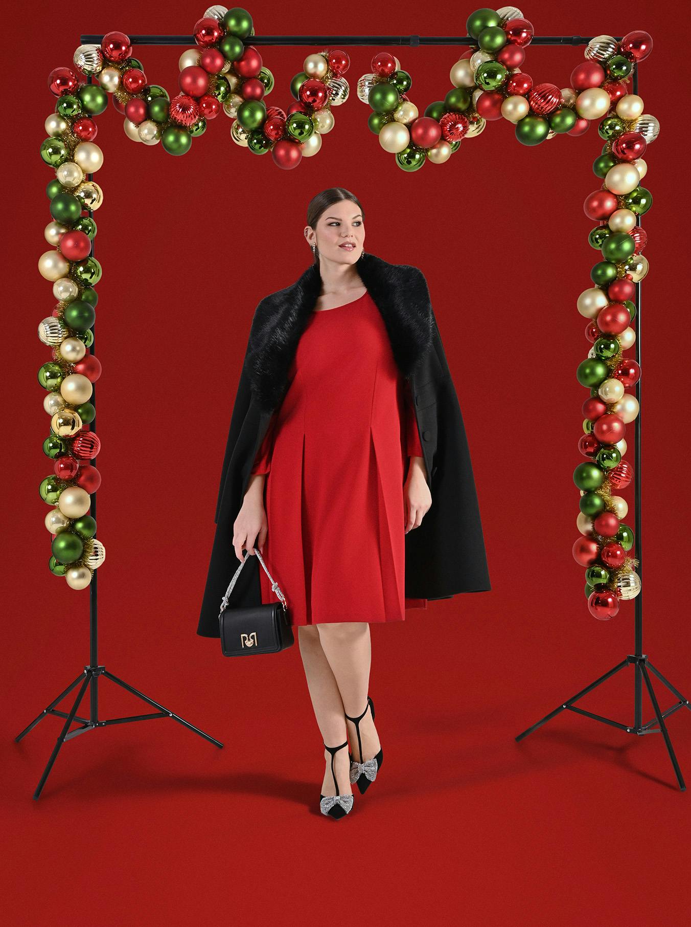 A woman in a red dress stands confidently against a red backdrop. She holds a small clutch and is framed by festive arches of red, green, and gold ornaments.