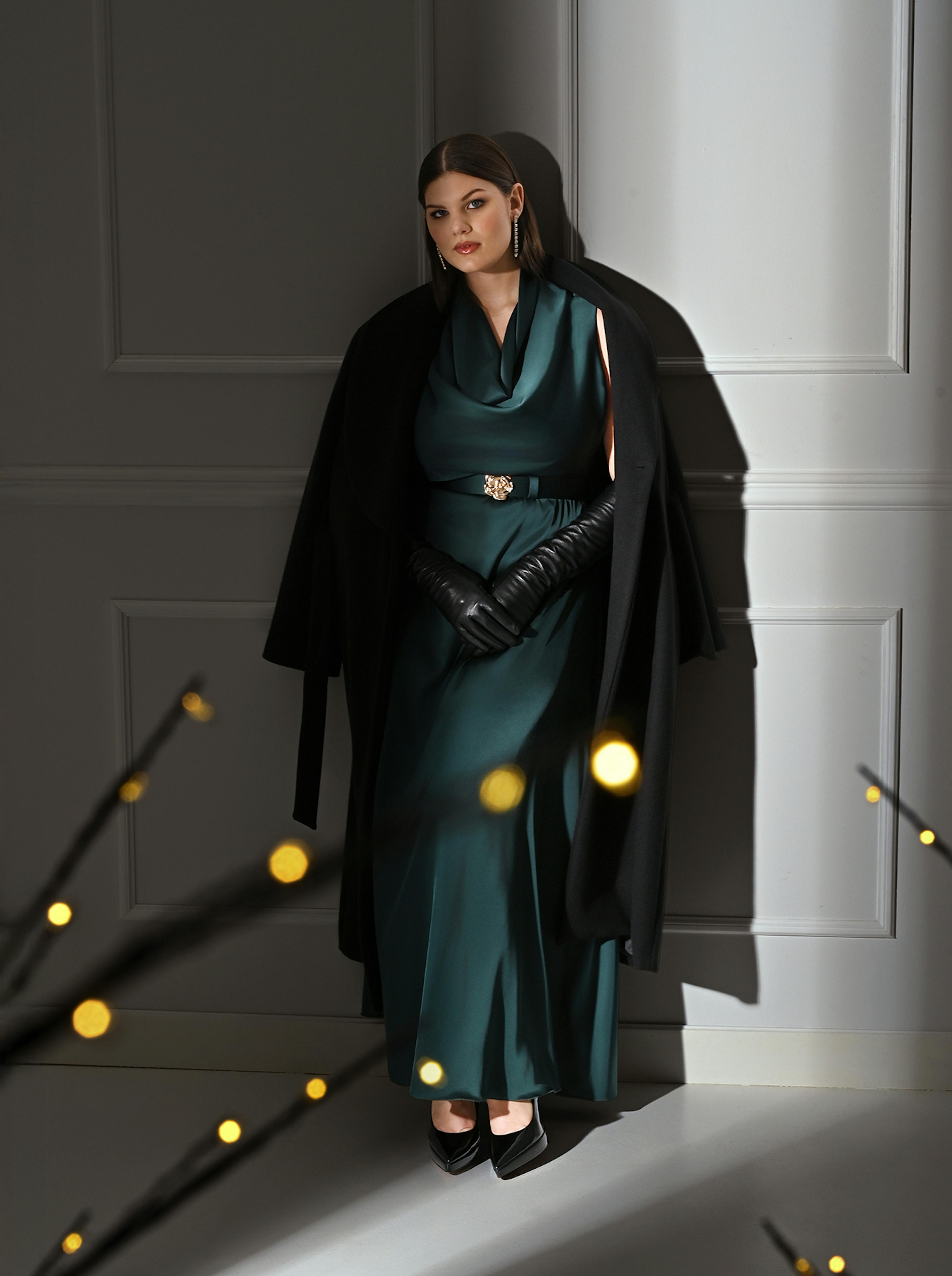 A woman in a teal dress and black coat leans against a wall. Her expression is confident, with elegant lighting creating a dramatic, sophisticated mood.