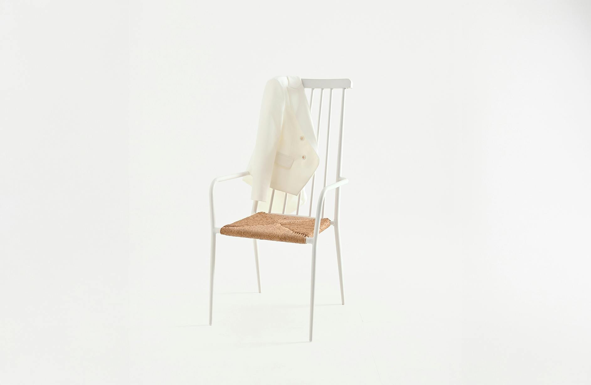 A minimalist white chair with a woven seat sits empty against a plain background. A white blazer is draped over its back, creating a serene, elegant atmosphere.