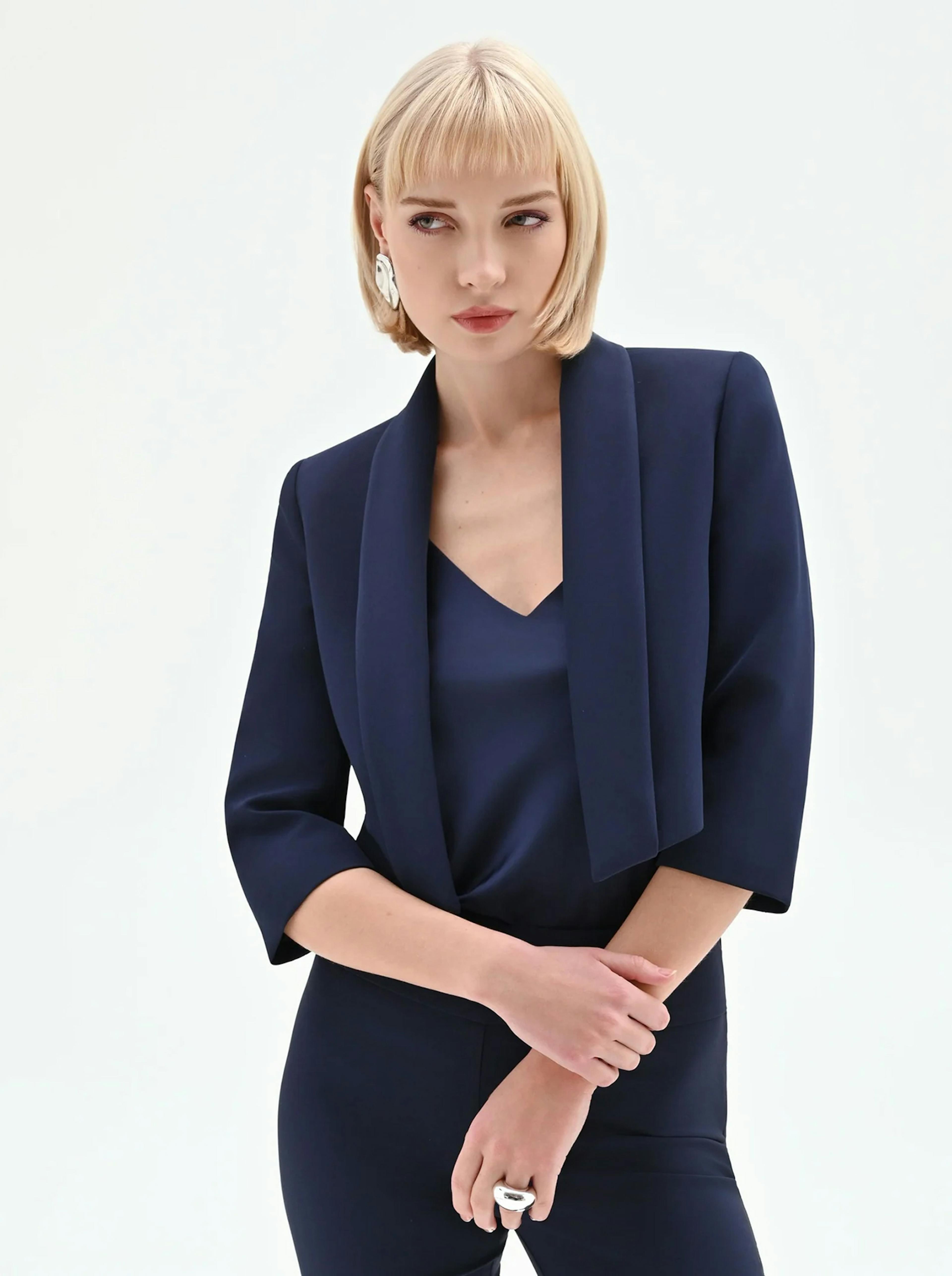 Woman with bob haircut poses confidently in a navy suit and blouse against a white background, conveying a professional and stylish aura.