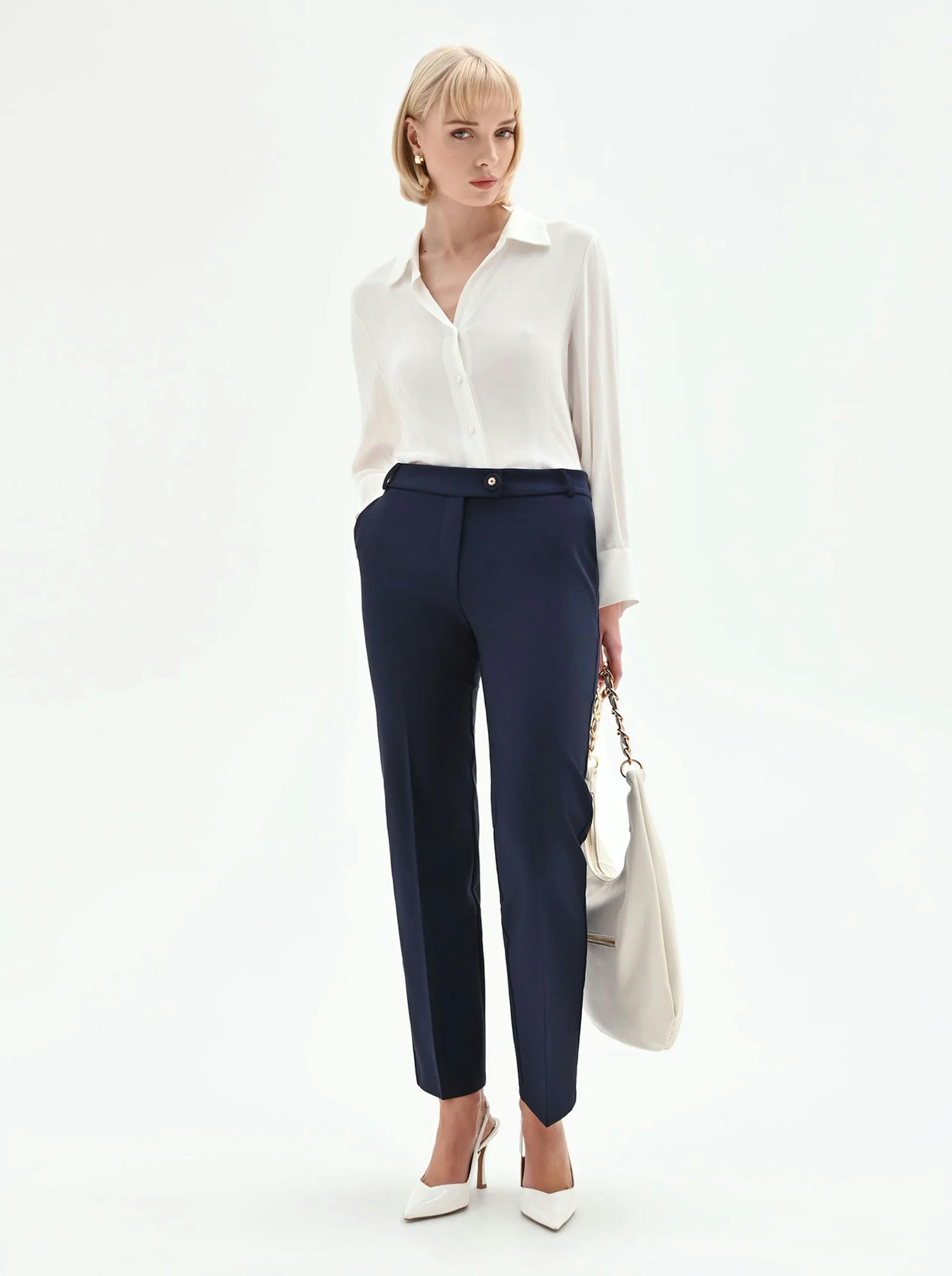 A woman stands poised in a white blouse and navy trousers, conveying a professional look. She holds a handbag and wears white heels, set against a plain background.