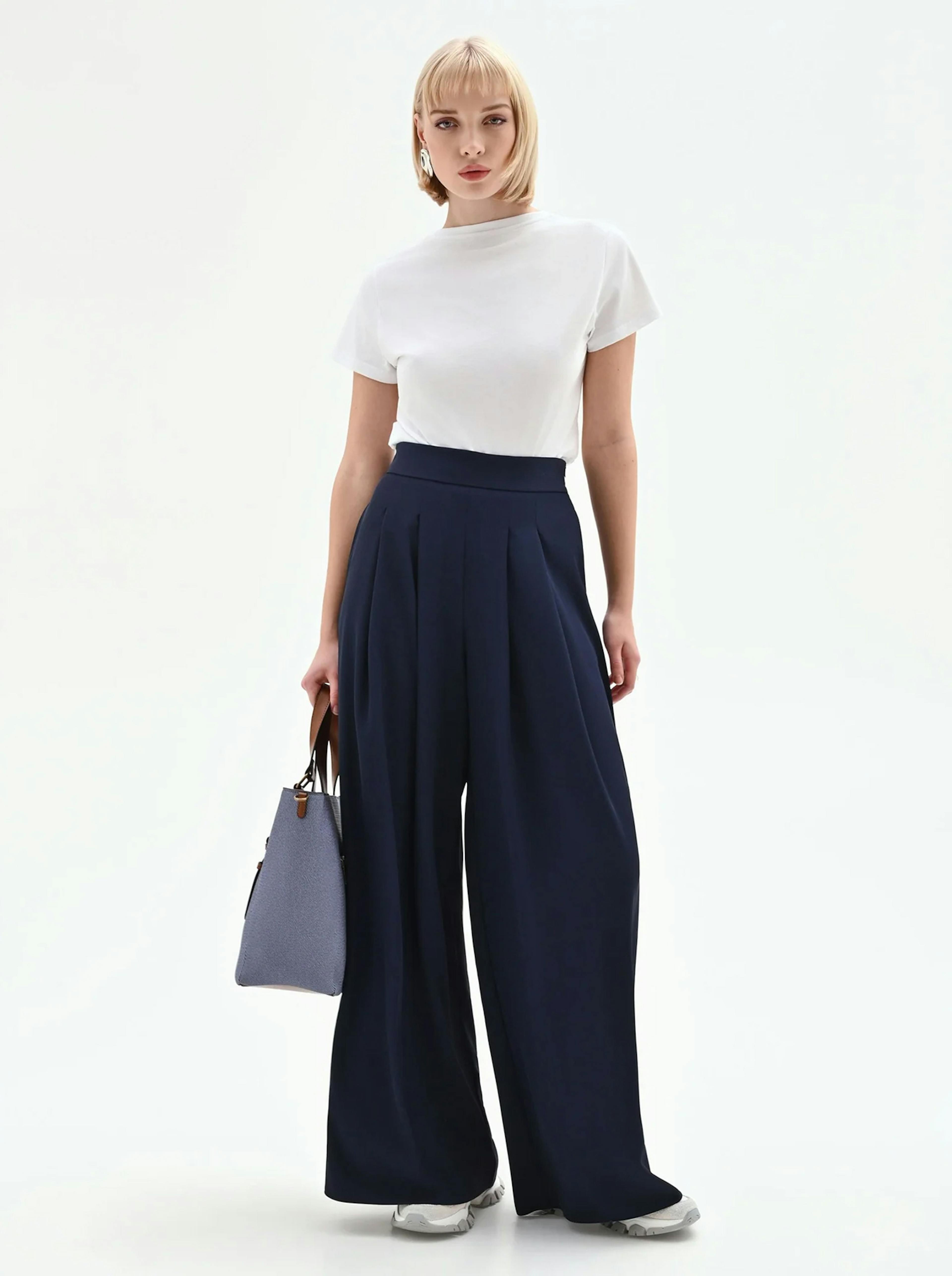 Woman in a white T-shirt and wide-leg navy trousers stands confidently against a plain background, holding a light blue handbag. Sophisticated and modern style.