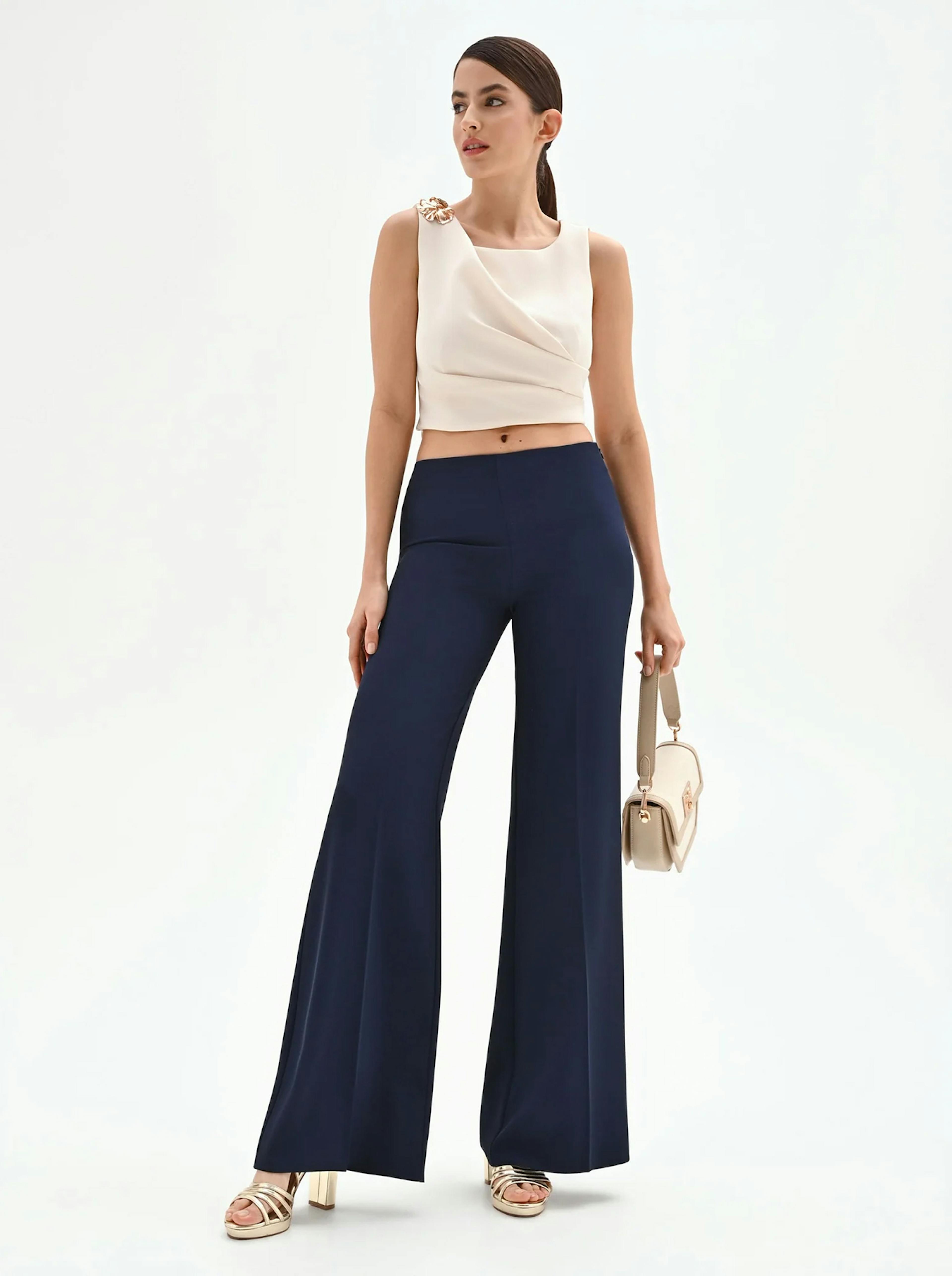 Woman in chic attire poses confidently. She wears a white sleeveless top and navy wide-leg pants, holding a beige handbag. Background is plain white.