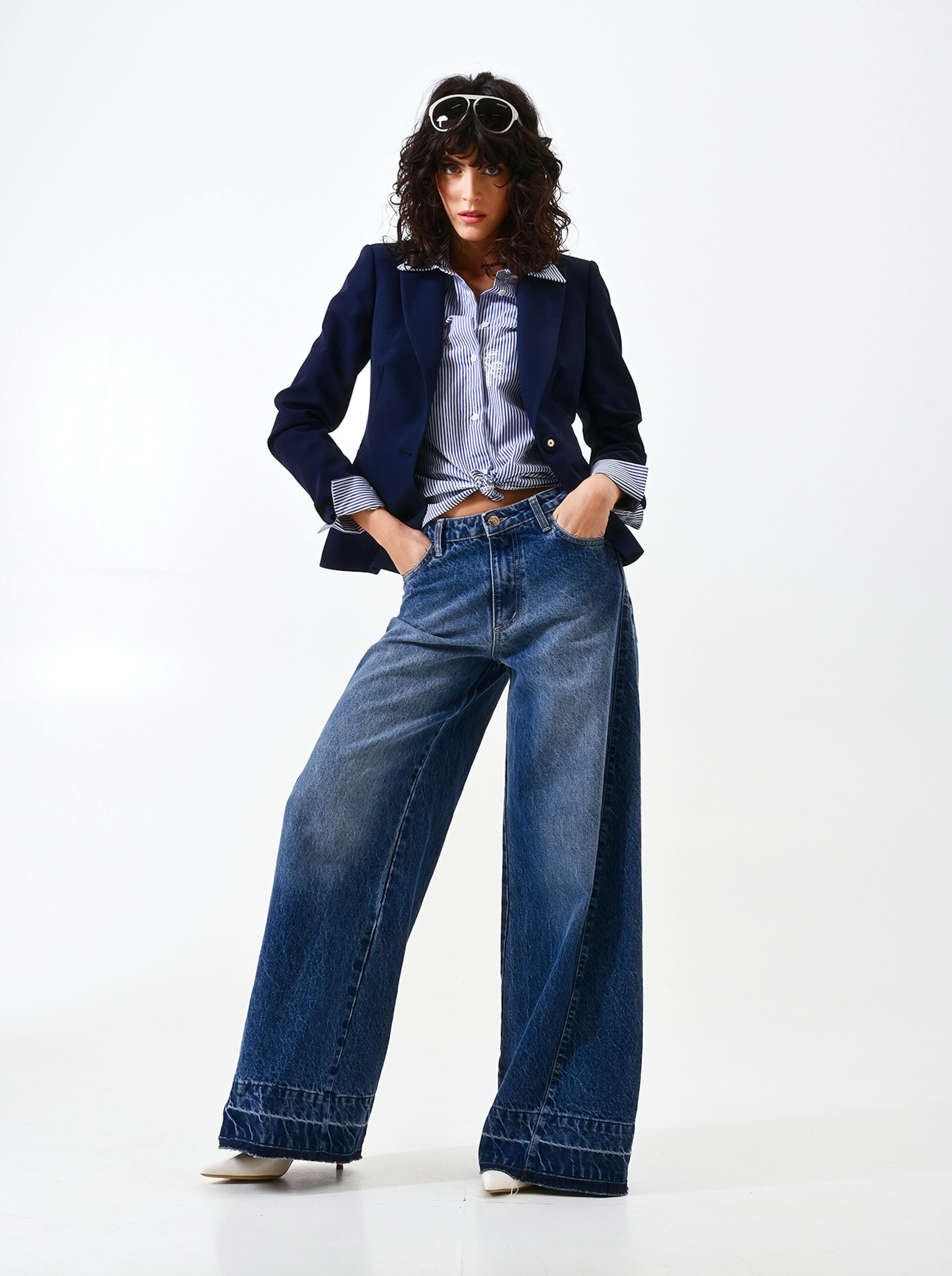 A person stands confidently in wide-legged jeans, a striped shirt, and a navy blazer. Sunglasses rest on their head, conveying a chic, relaxed style.