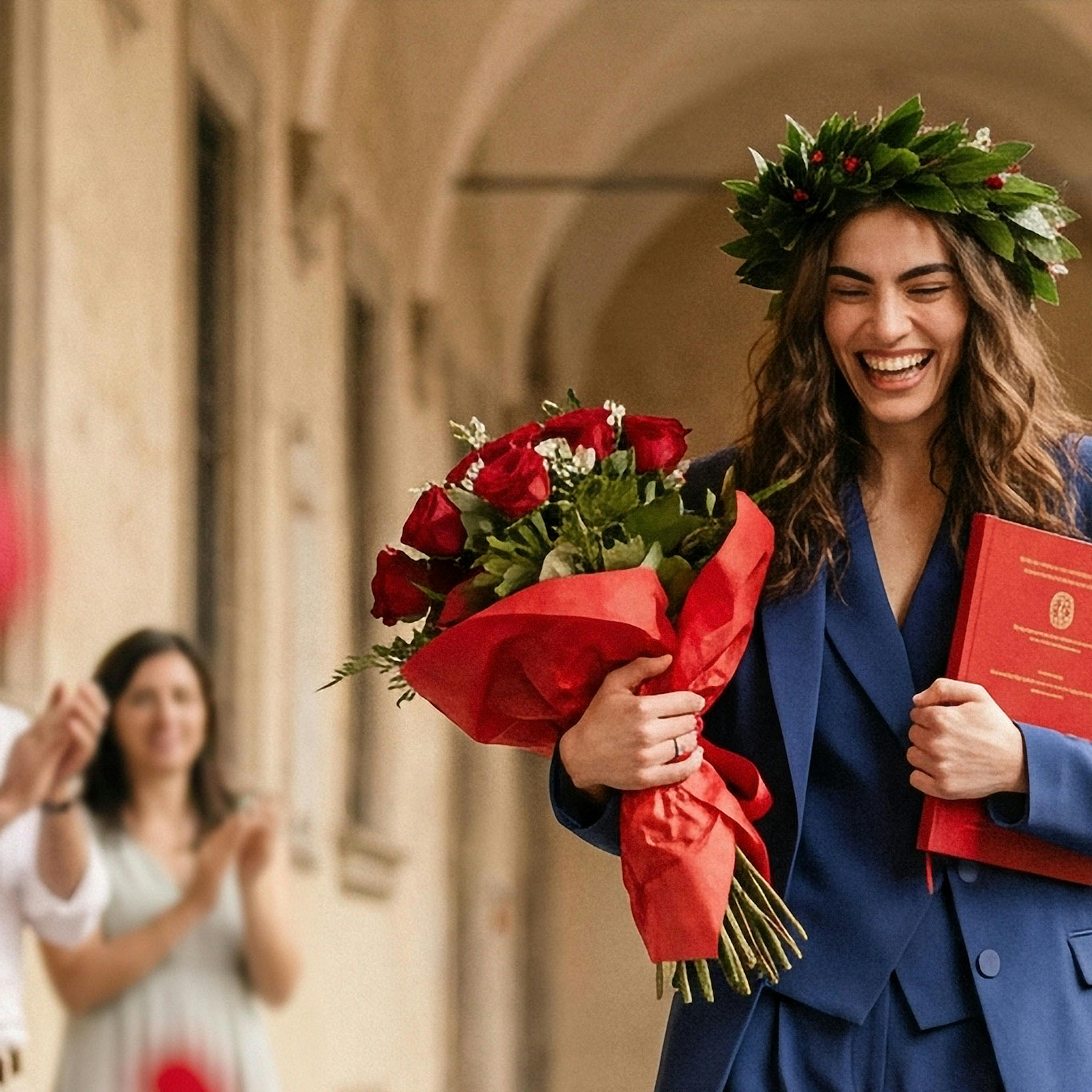 A joyful woman in a blue suit holds a red diploma and roses, with a laurel wreath on her head. Red petals are thrown, and people clap in an arched hallway.