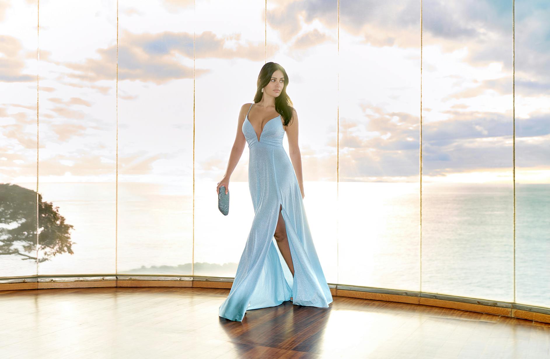 A woman in a flowing light blue gown stands by a large window at sunset, holding a clutch. The ocean and sky create a serene backdrop.