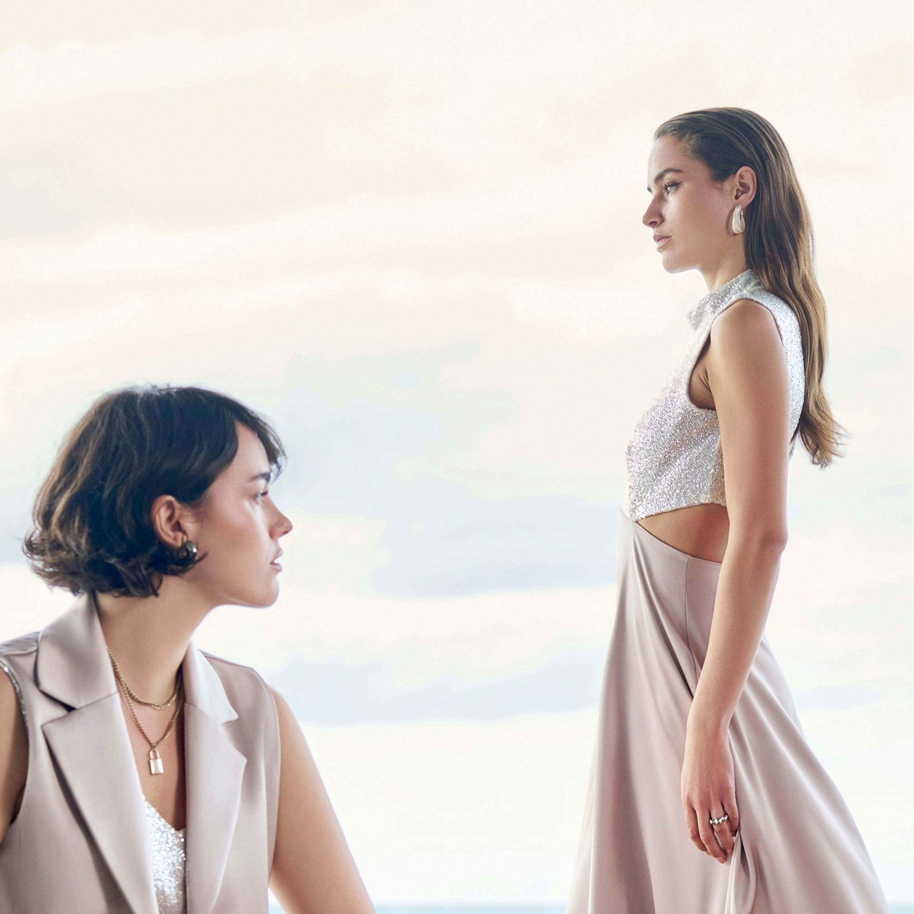 Two women in elegant dresses are posed by a large window with a cloudy sky behind them. One sits gazing forward; the other stands in profile.