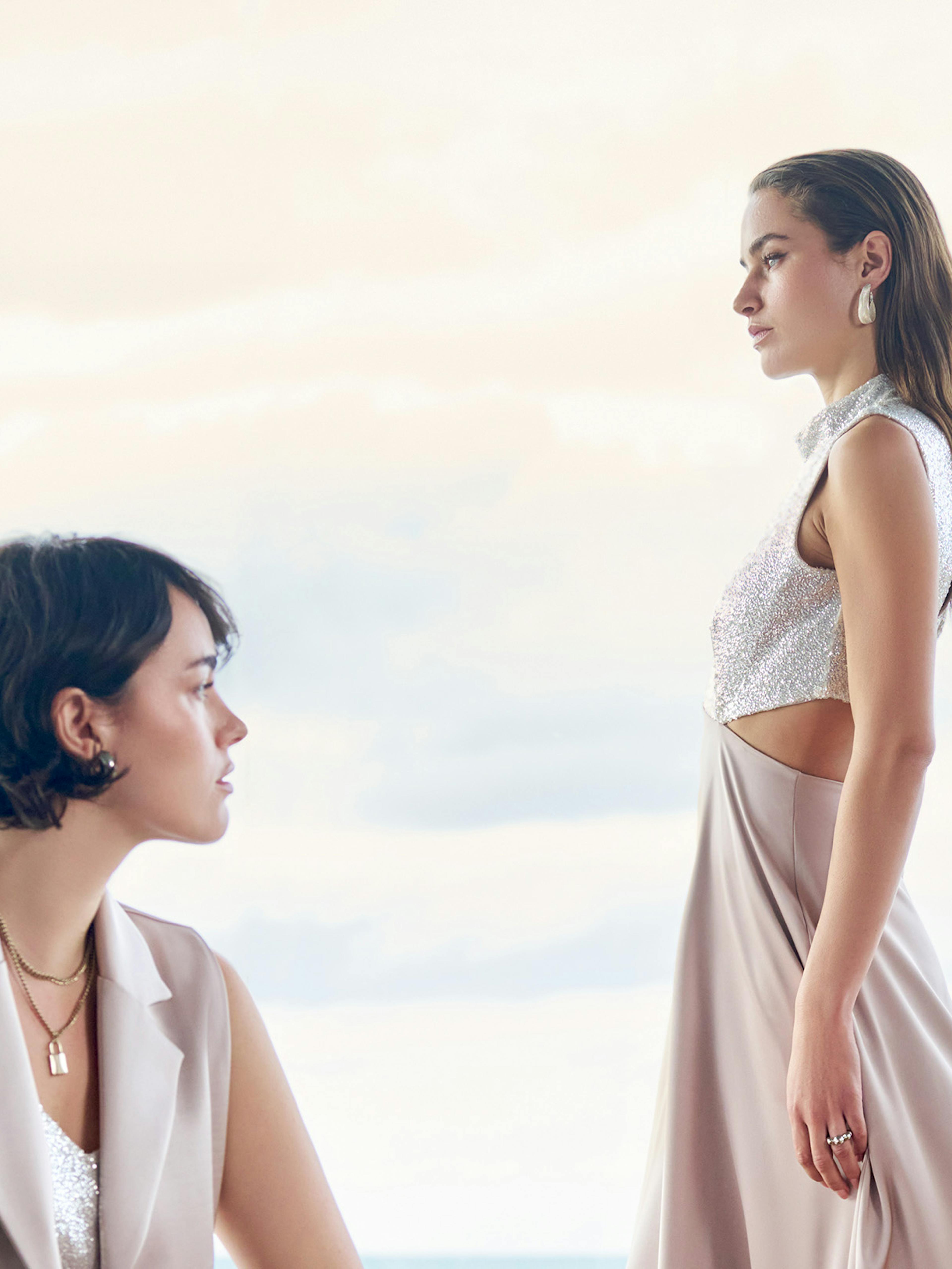 Two women in elegant dresses are posed by a large window with a cloudy sky behind them. One sits gazing forward; the other stands in profile.
